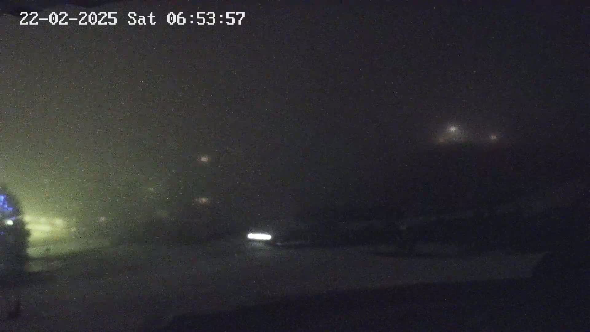 A nighttime, foggy scene shows a vehicle's headlights illuminating a dark road with faintly visible lights in the distance.