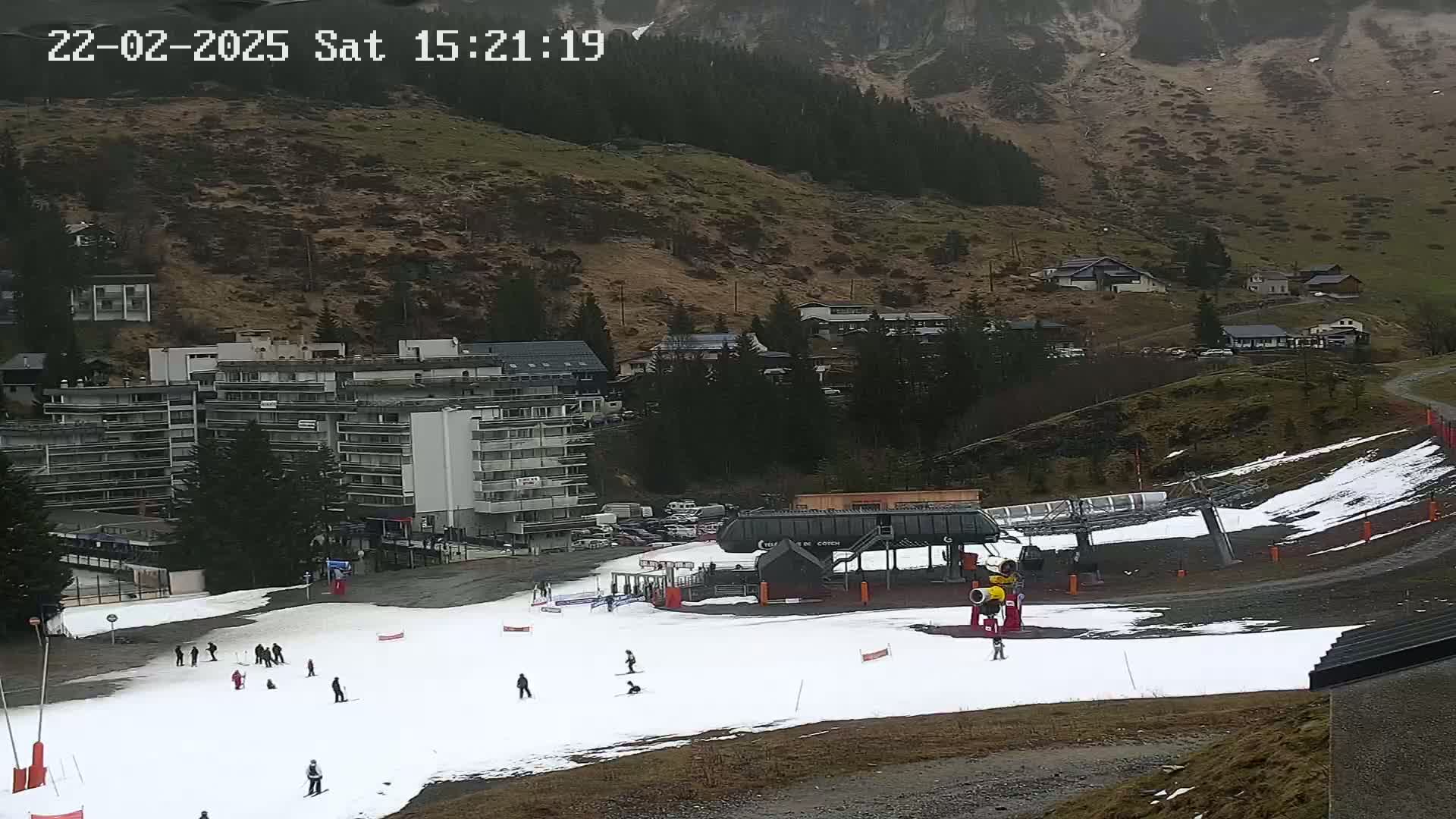 A ski slope with a few skiers, a ski lift, and a large apartment building is situated on a mountainside with overcast weather.