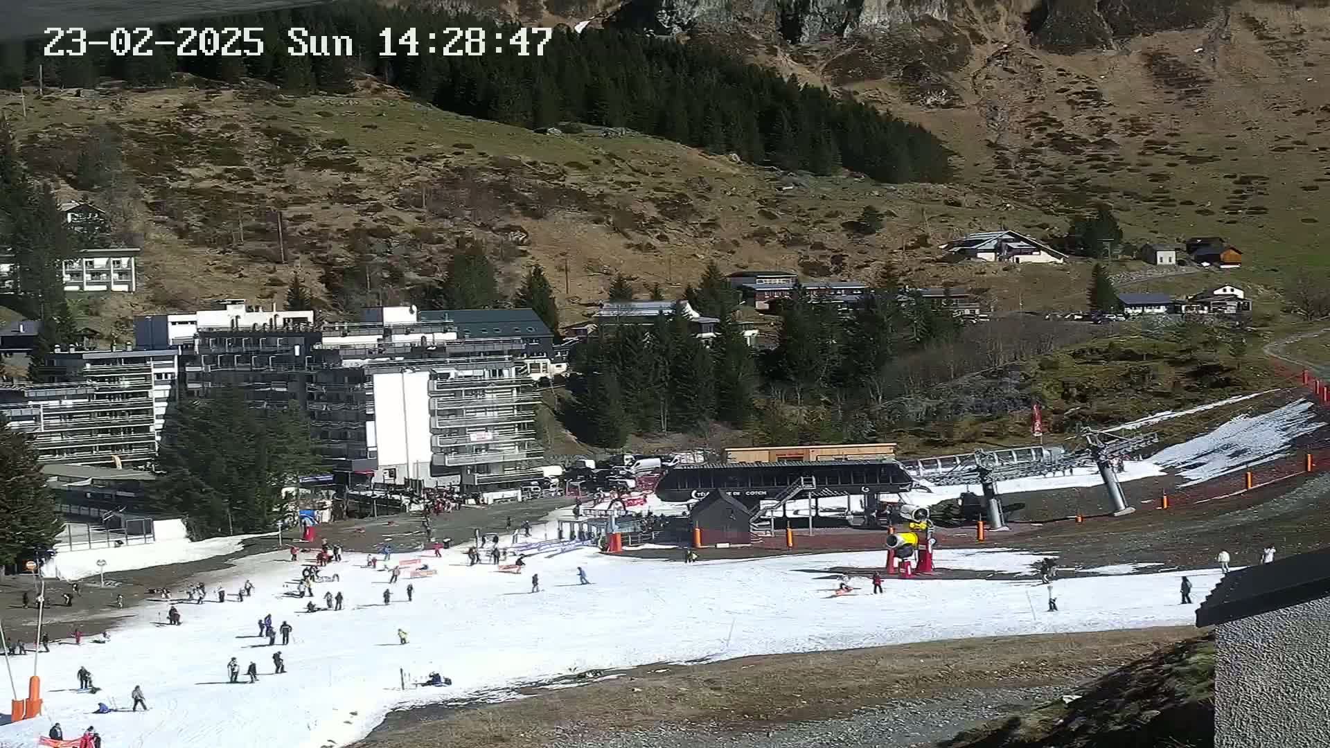 A ski slope with a chairlift and several buildings in a mountainous area on a sunny day with patches of snow.