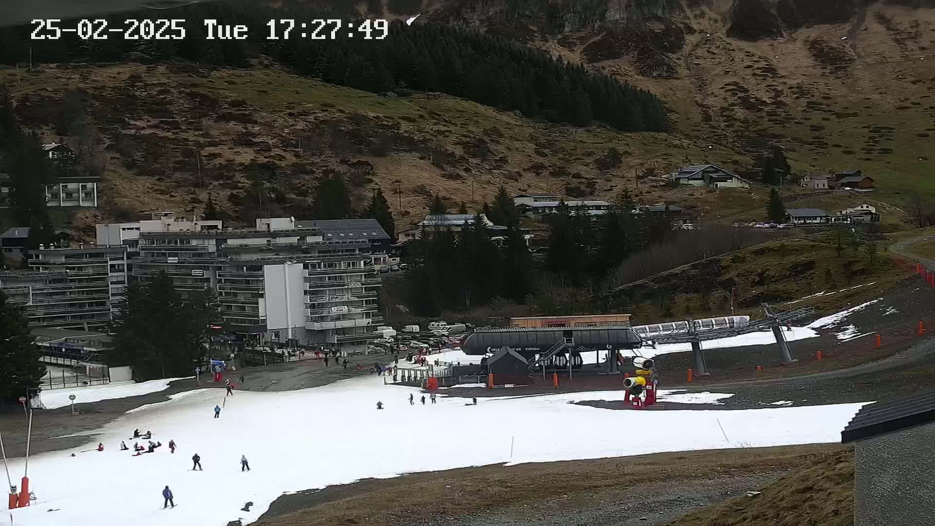 A ski slope with a chairlift and a large apartment building is visible, with a few people skiing and snowboarding on the snow-covered ground, and overcast weather conditions.
