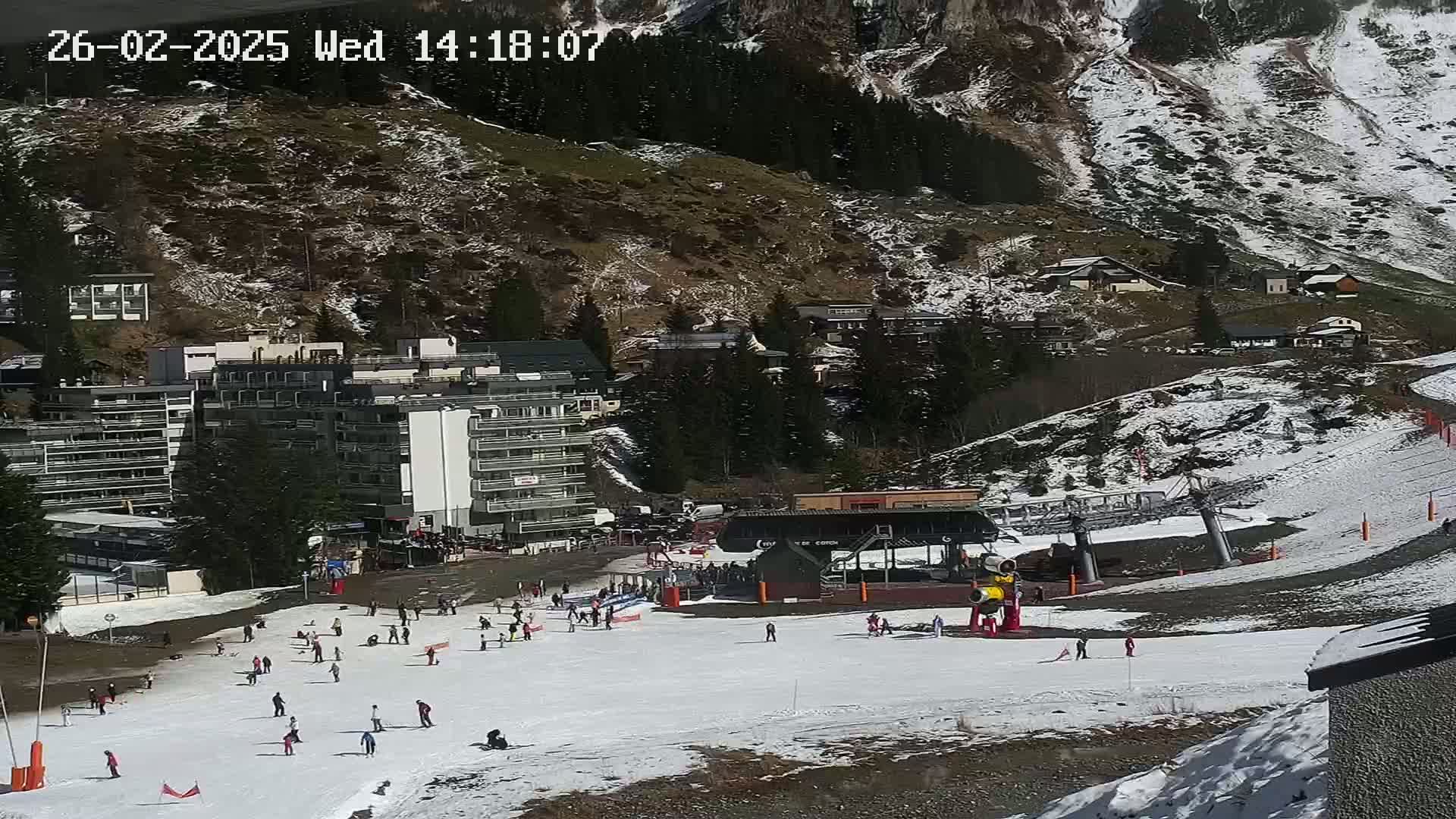 A ski slope with scattered skiers and a ski lift, situated in a mountainous area with patchy snow cover and sunny weather.