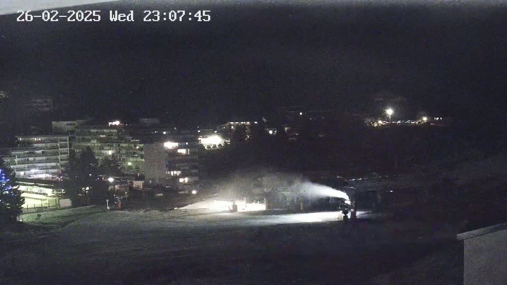At night, a snow groomer makes snow in a snowy area near several buildings under a dark sky.