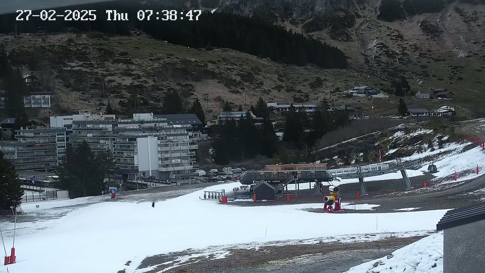 A snow-covered ski slope with a chairlift and a large apartment building sits below a mountainside with sparse vegetation and some residential buildings, under a mostly cloudy sky.