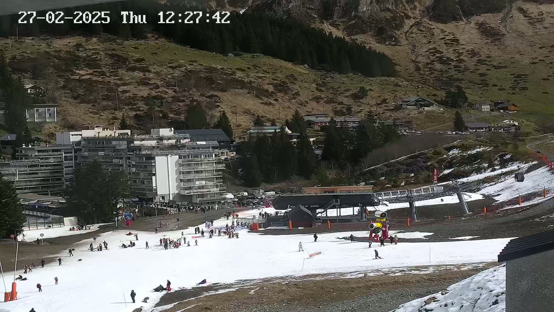 A ski slope with patches of snow, a ski lift, several people, and a large building situated against a mountainside with sparse snow and evergreen trees.