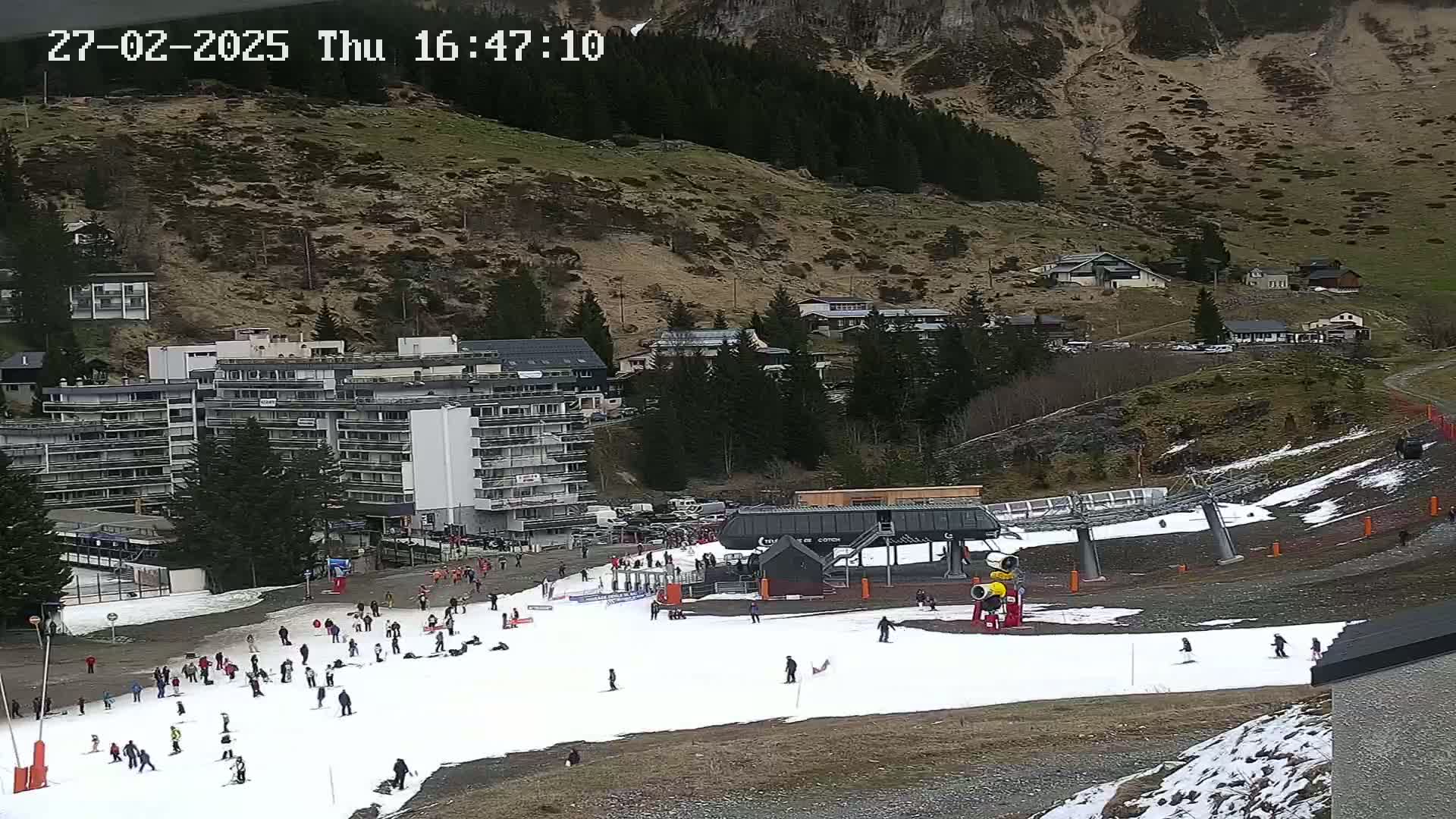 A ski slope with a few skiers and snowboarders, a ski lift, and several buildings in a mountainous area with partly cloudy skies.