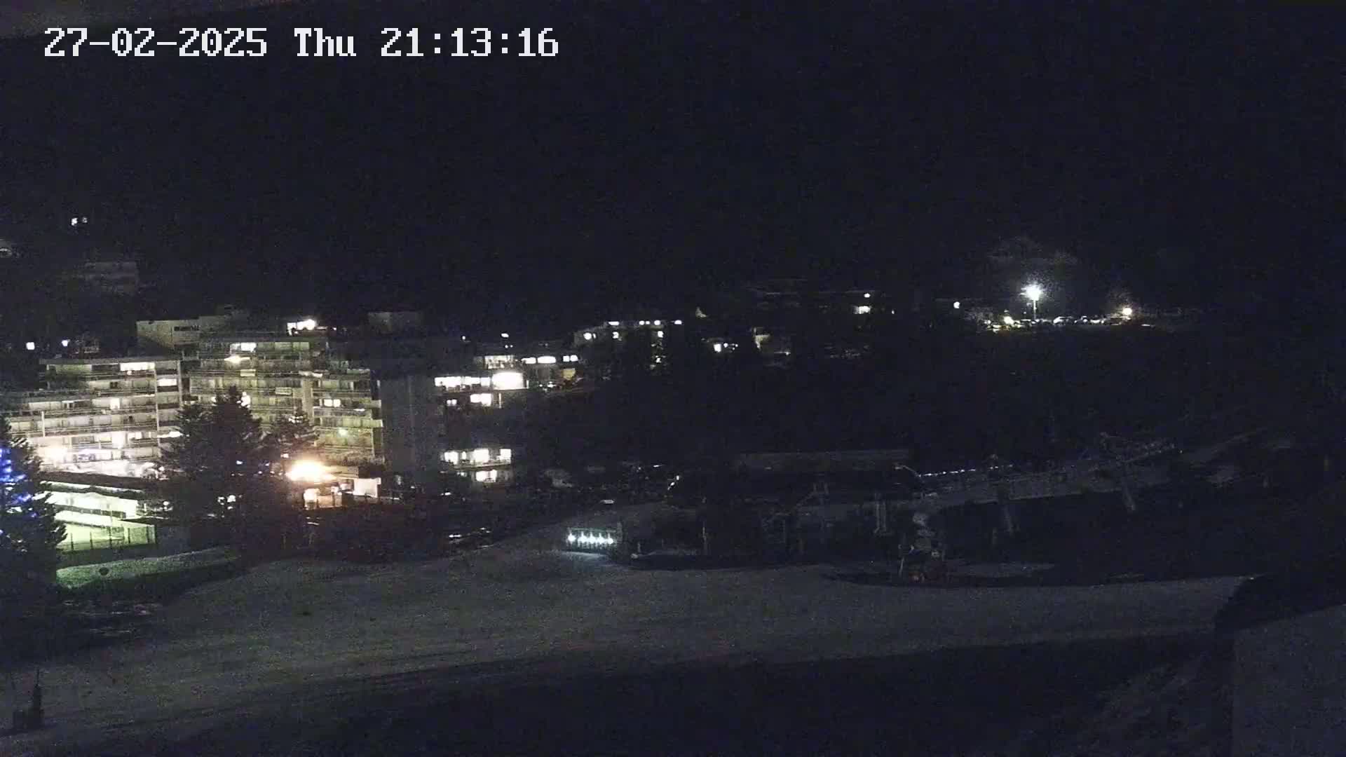 A nighttime view of a snow-covered area with several illuminated buildings in the background under a dark sky.