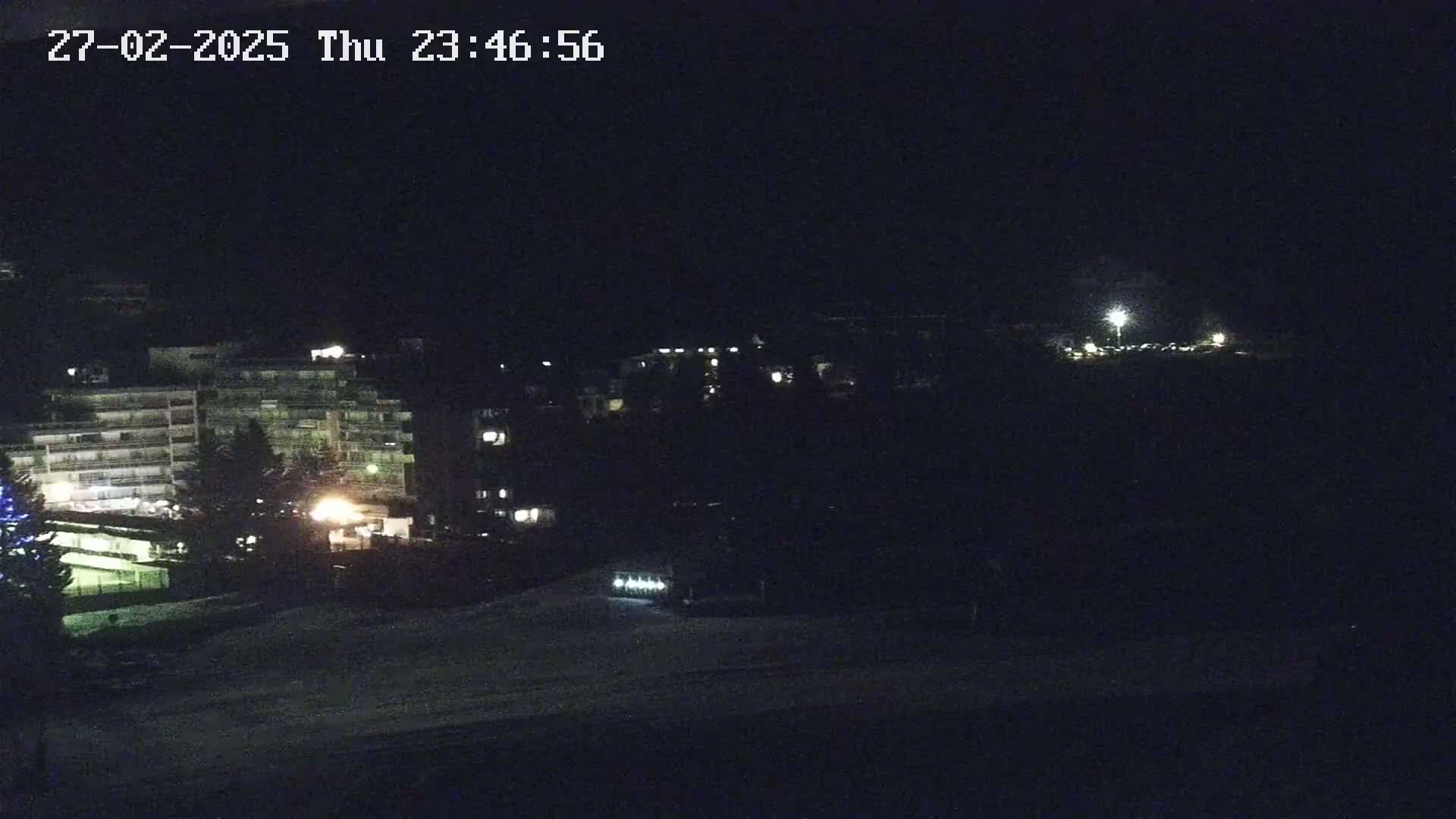 A nighttime, snow-covered scene shows several multi-story buildings with some lights on, a parking area with a few lights, and some trees.