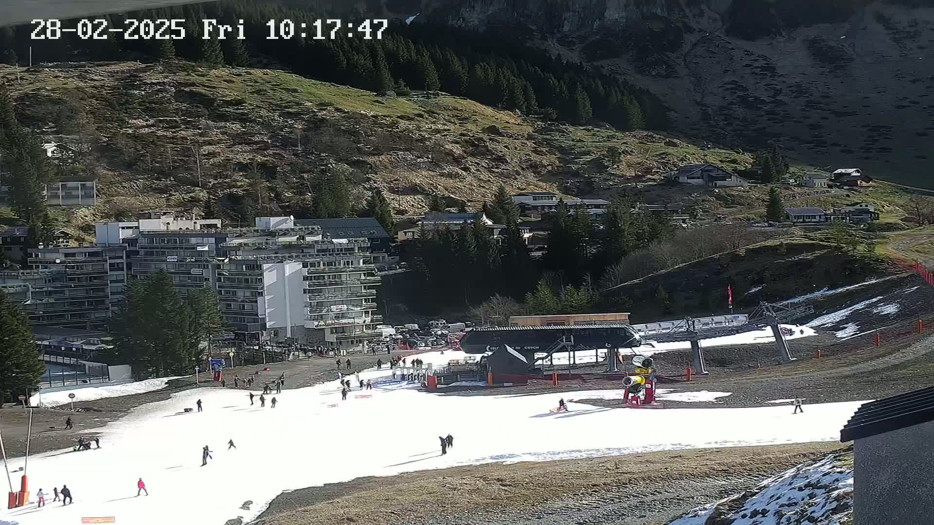 A ski slope with a chairlift and several people skiing or snowboarding, situated near a mountainside village under sunny conditions.