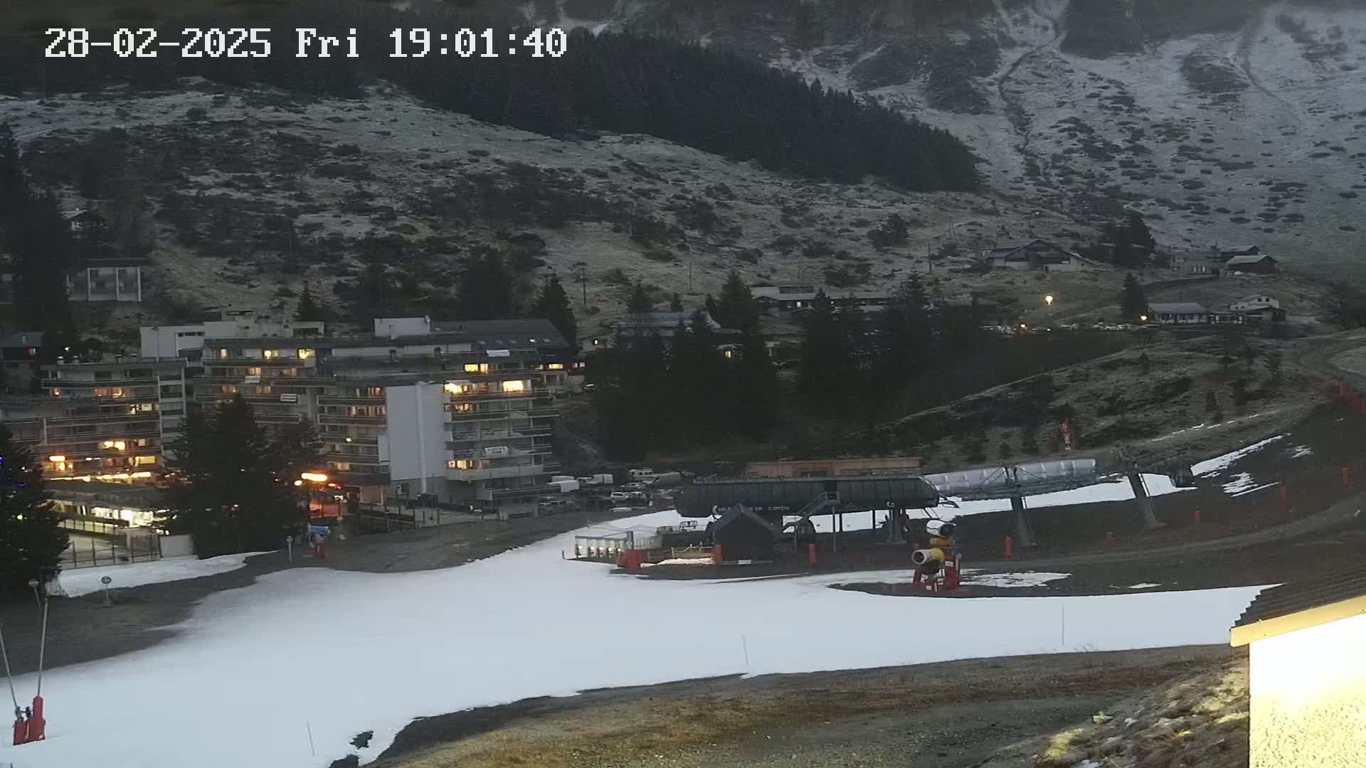 A snow-covered ski slope at dusk, with a ski lift and apartment buildings illuminated against a mountainside dusted with snow.