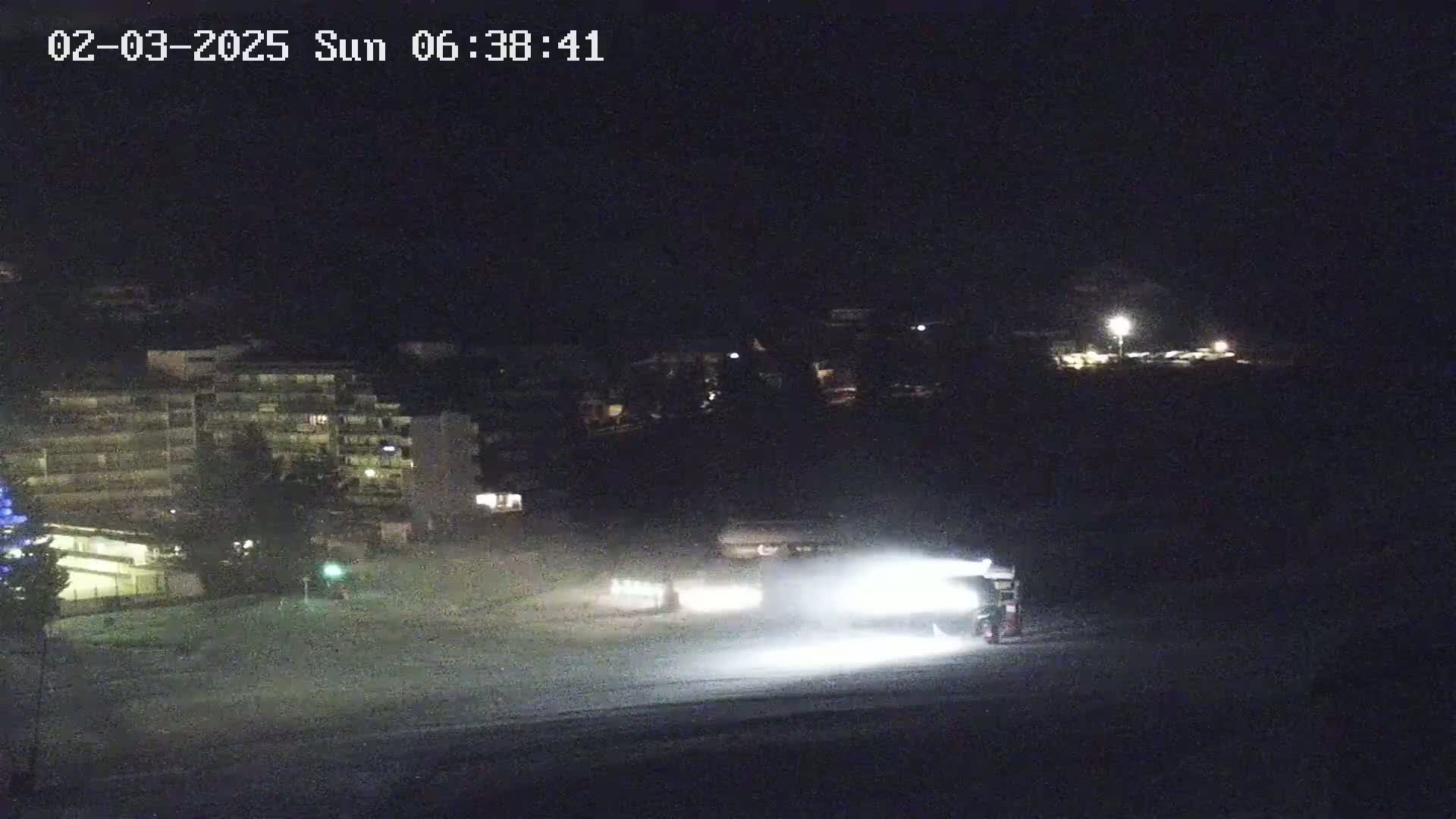 At night, snow groomers illuminate a snow-covered area in front of a building complex under a dark sky.