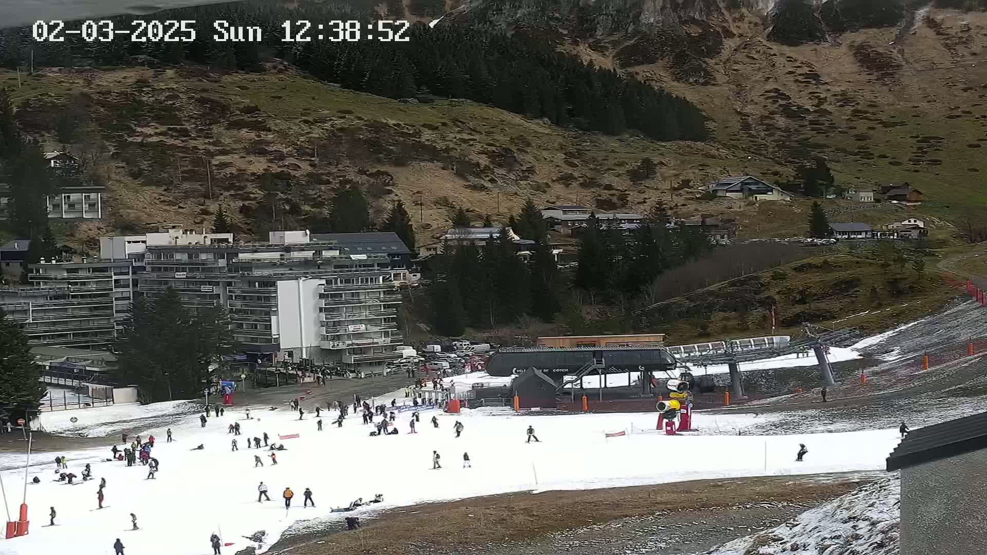 A ski slope with scattered skiers and a chairlift, situated below a mountainside with buildings and trees, on a partly cloudy day.