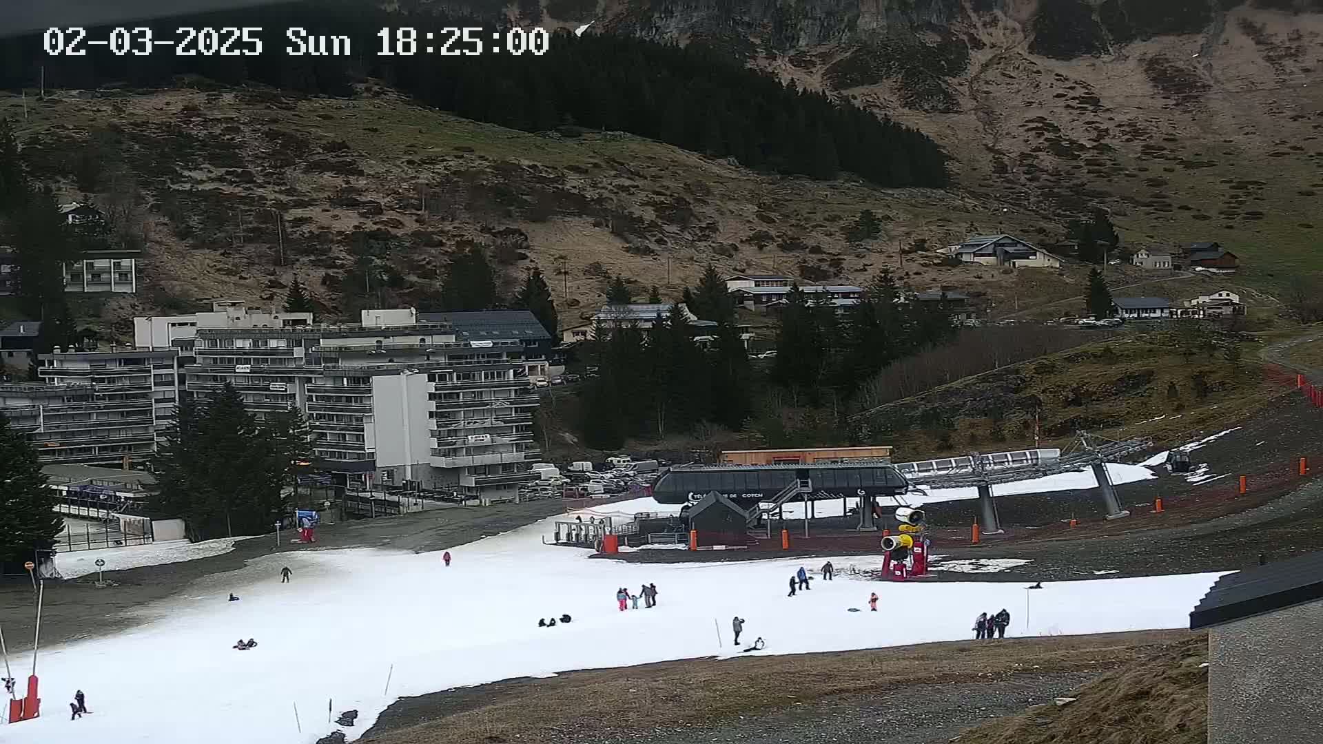 A ski lift and a snow-covered slope with several people are situated below a mountainside village on an overcast day.