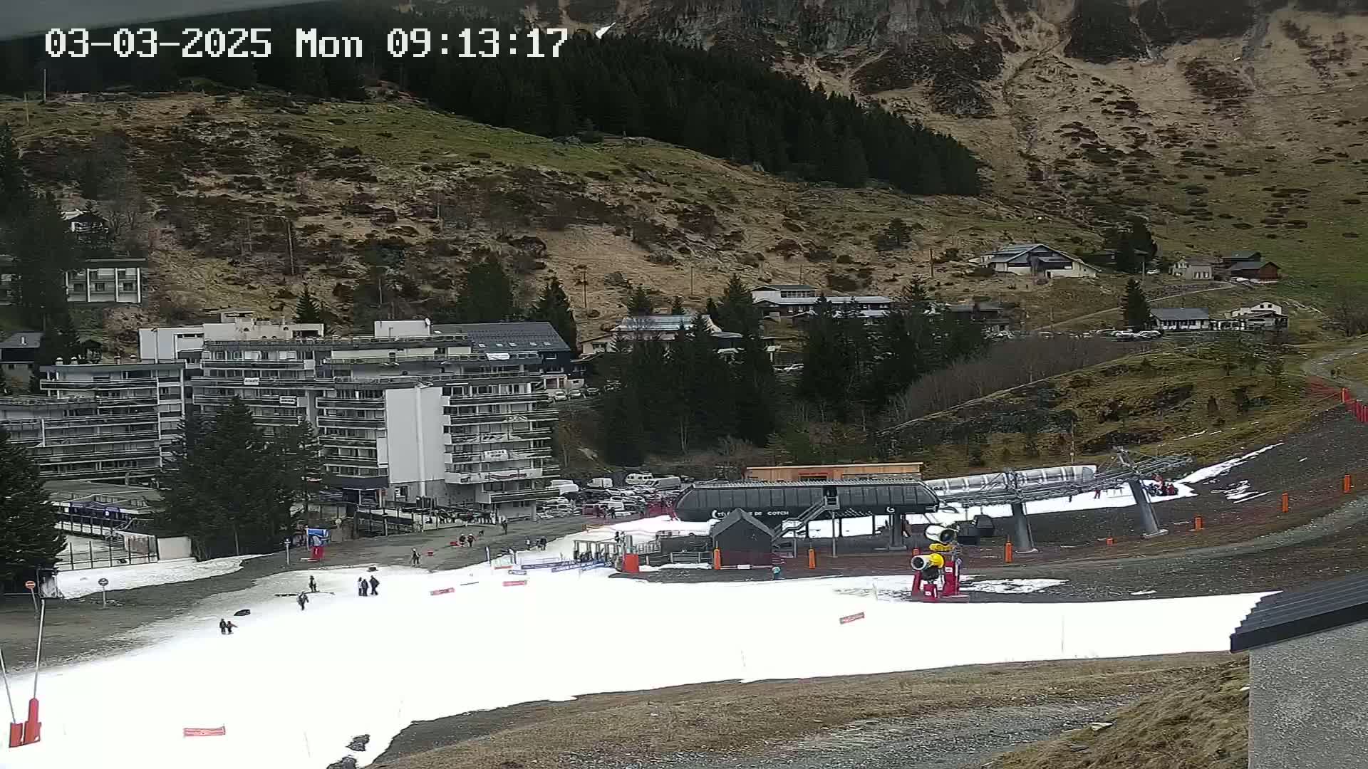 A ski slope with a chairlift and a few people on it, a multi-story building, and several other buildings on a hillside, under an overcast sky.