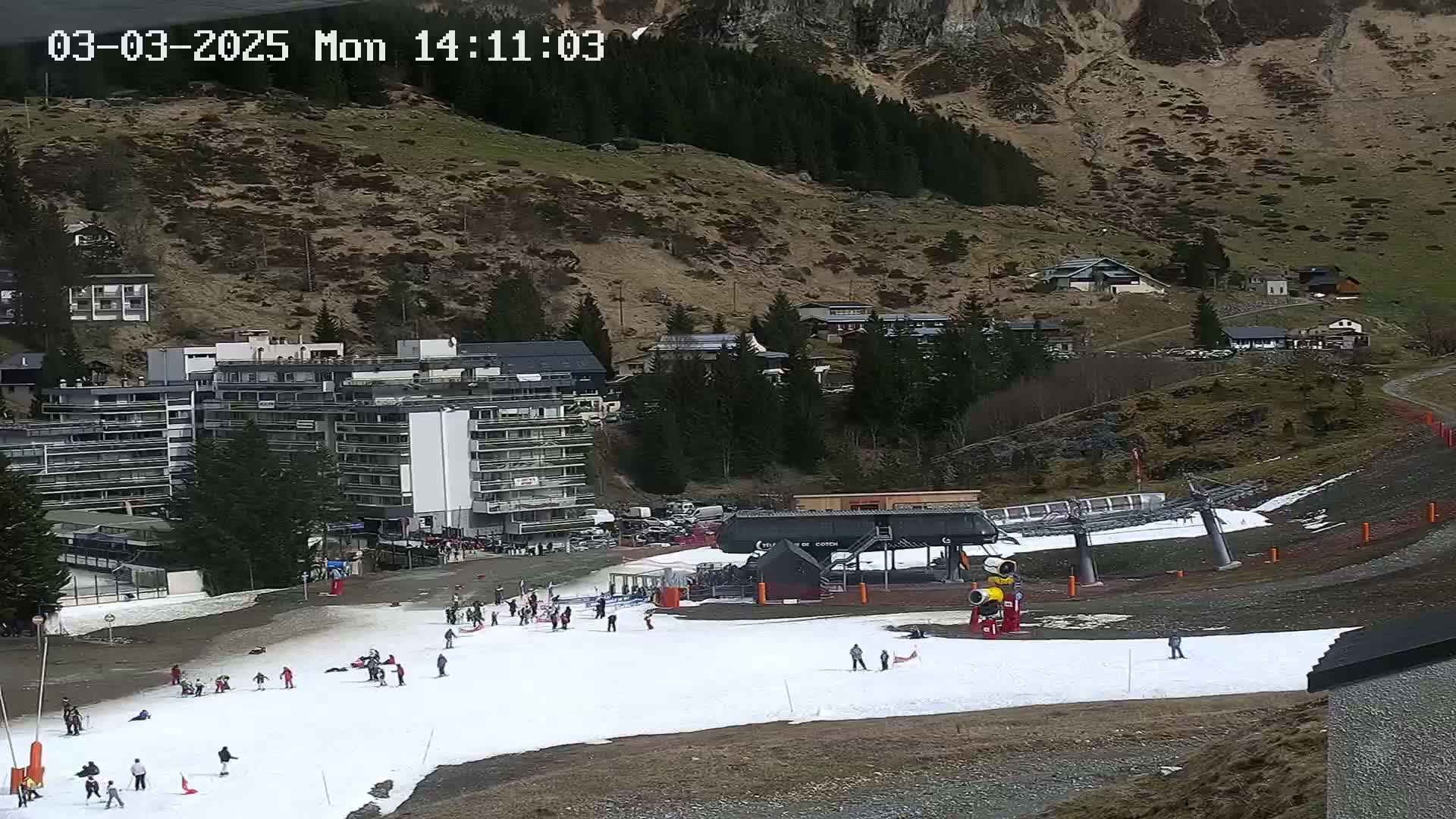 A ski slope with sparse snow cover, a chairlift, several buildings, and numerous people are visible against a mountainous backdrop under a seemingly clear sky.