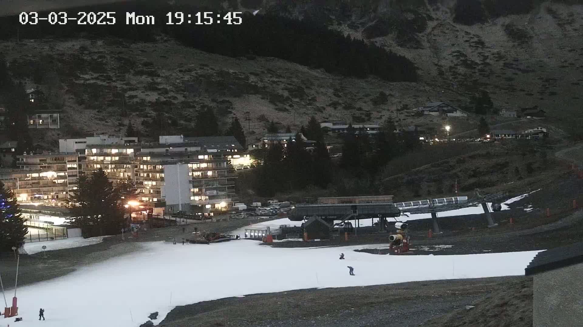 A snow-covered slope at dusk, with a ski lift and several buildings illuminated in the background, situated on a mountainside.