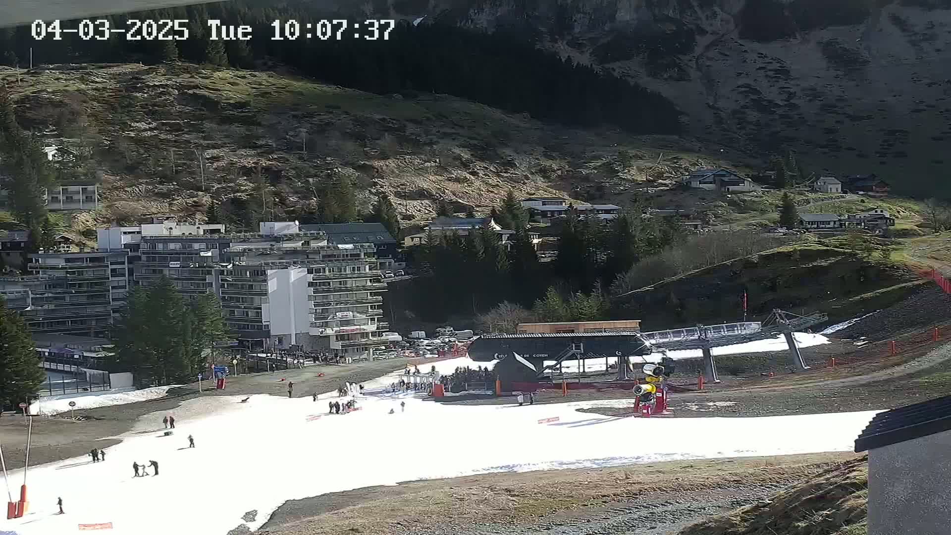 A snow-covered ski slope with a ski lift and several people, situated below a mountainside village on a sunny day.