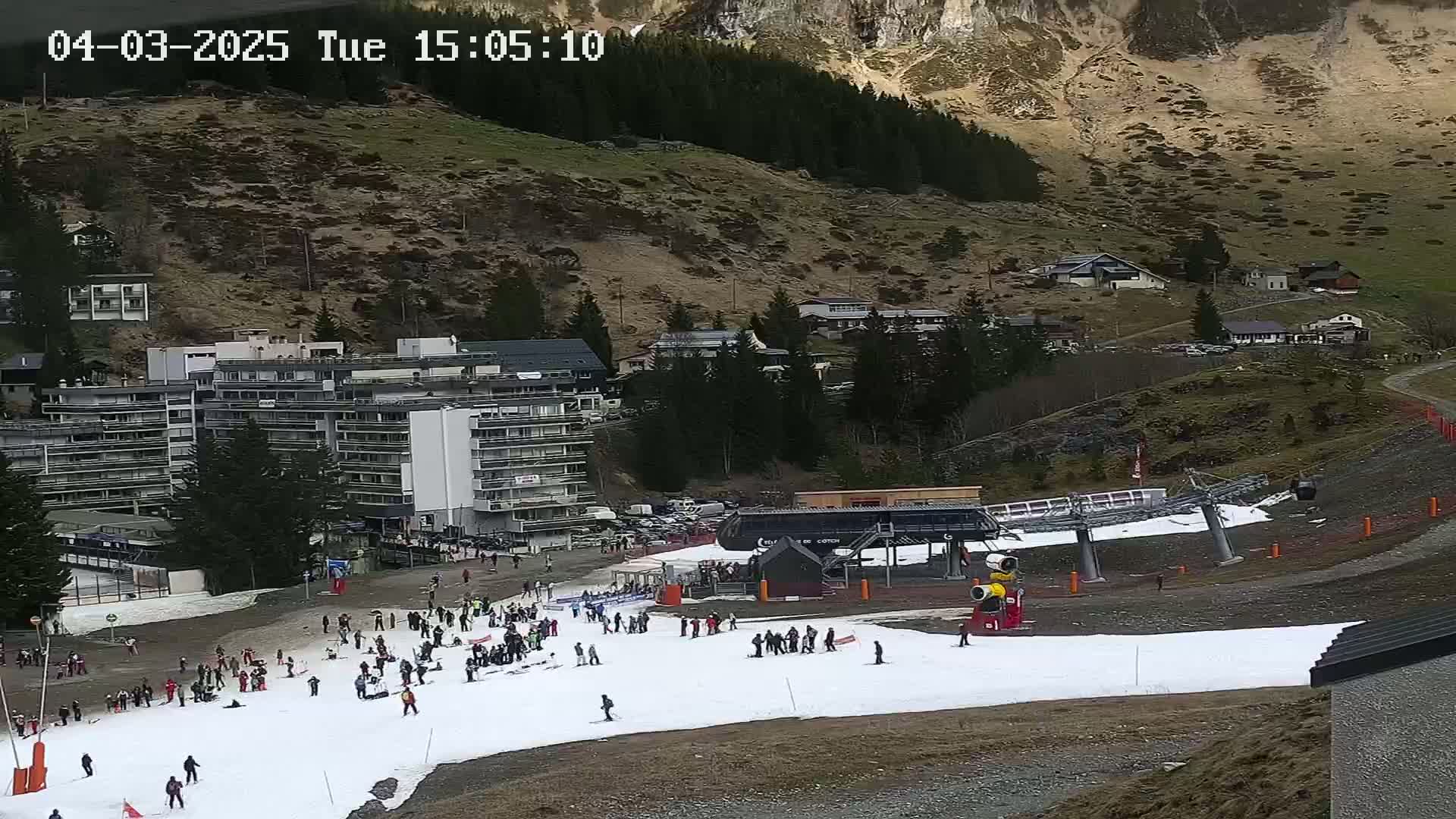 A ski slope with a small number of skiers and snowboarders, a chairlift, and a large apartment building are situated on a mountainside under sunny conditions.