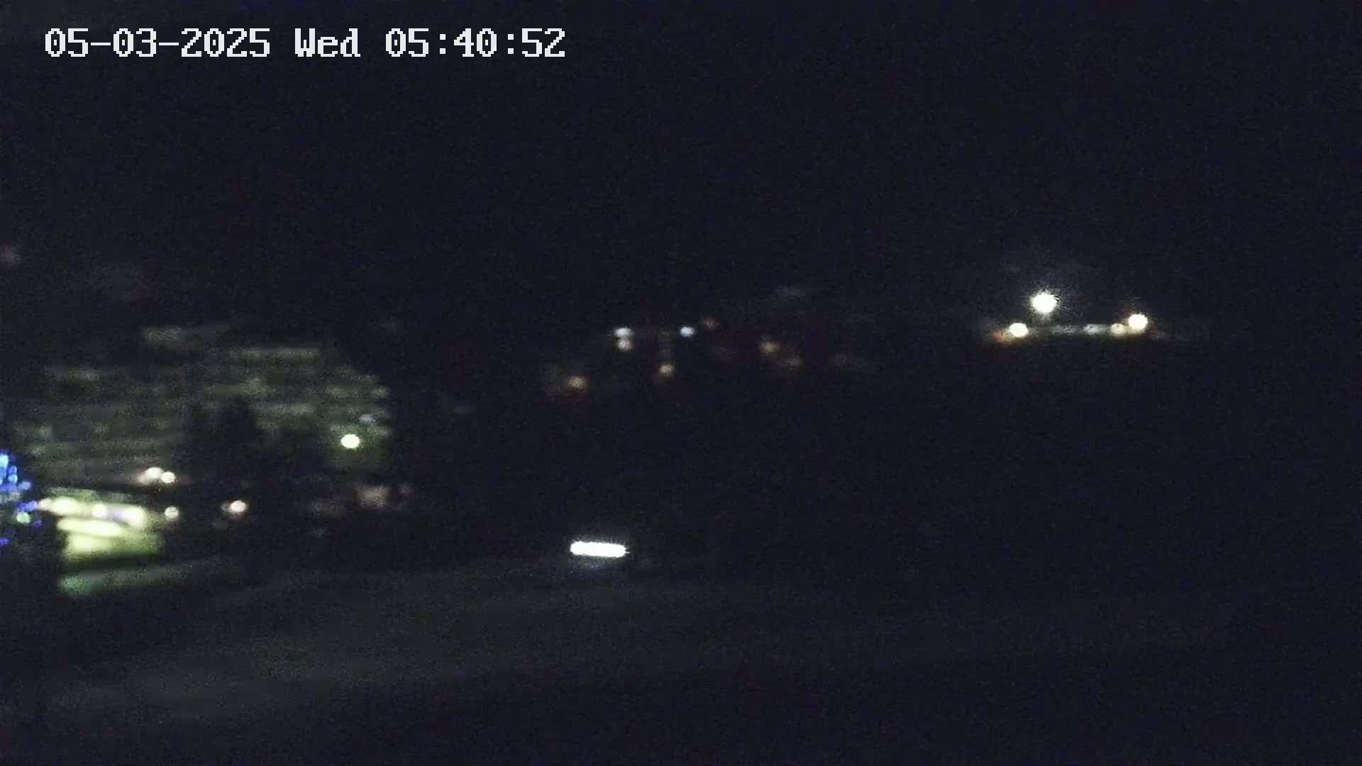 A nighttime long shot shows a blurry view of distant buildings and a vehicle's headlights on a dark road.
