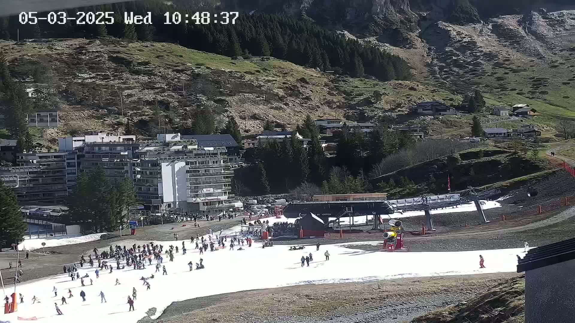 A ski area with a snow-covered slope, a ski lift, several buildings, and many people, situated on a mountainside under a sunny sky.