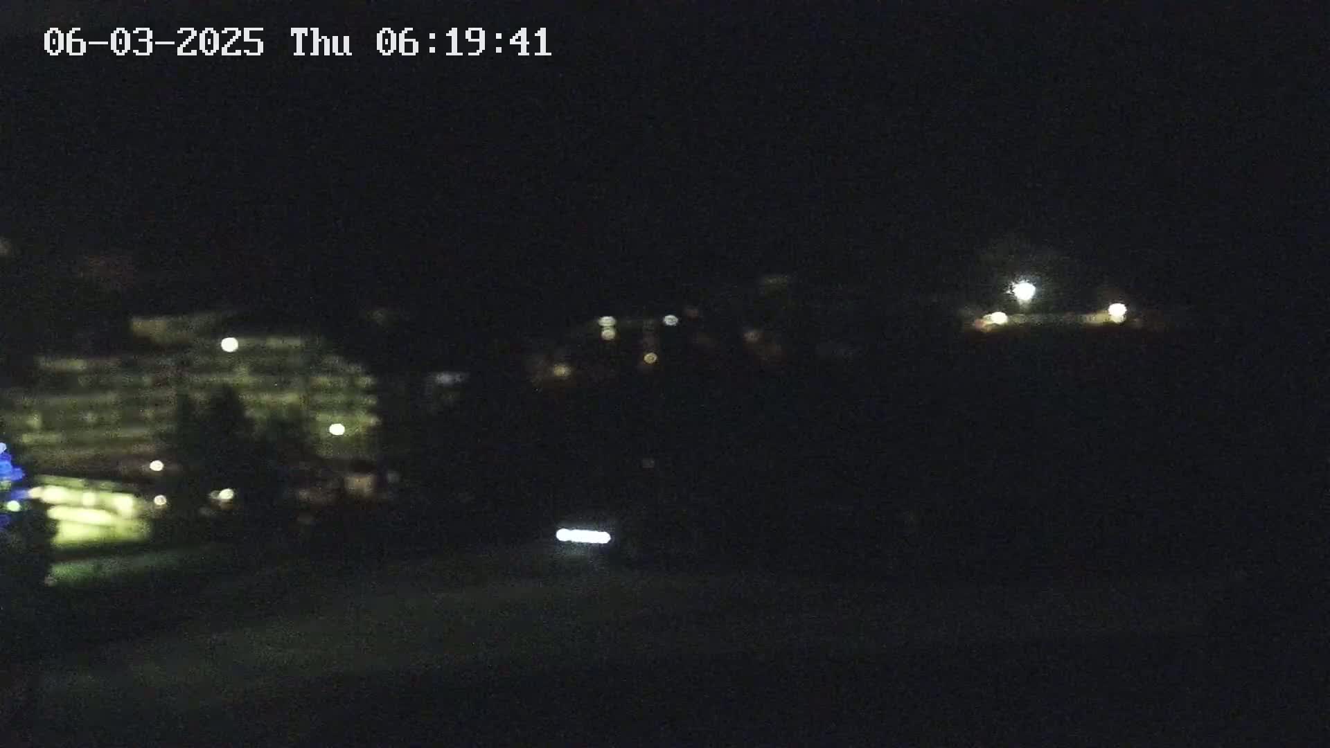 A nighttime, blurry view of a building with lights on, other distant lights, and a vehicle's headlights in the foreground.