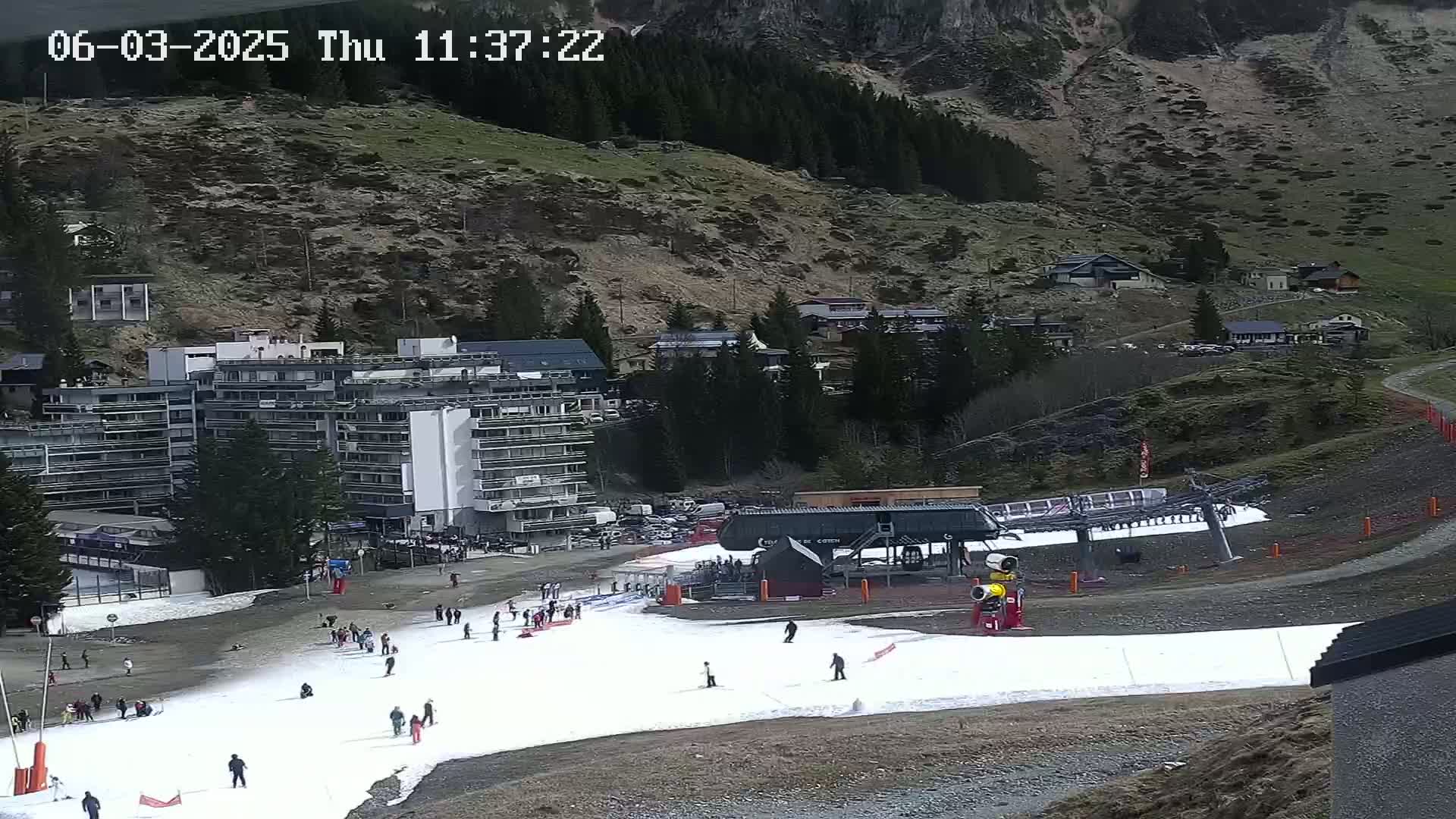 A ski slope with a few skiers and a chairlift is situated below a large apartment building and a mountainside covered in sparse vegetation, under a cloudy sky.