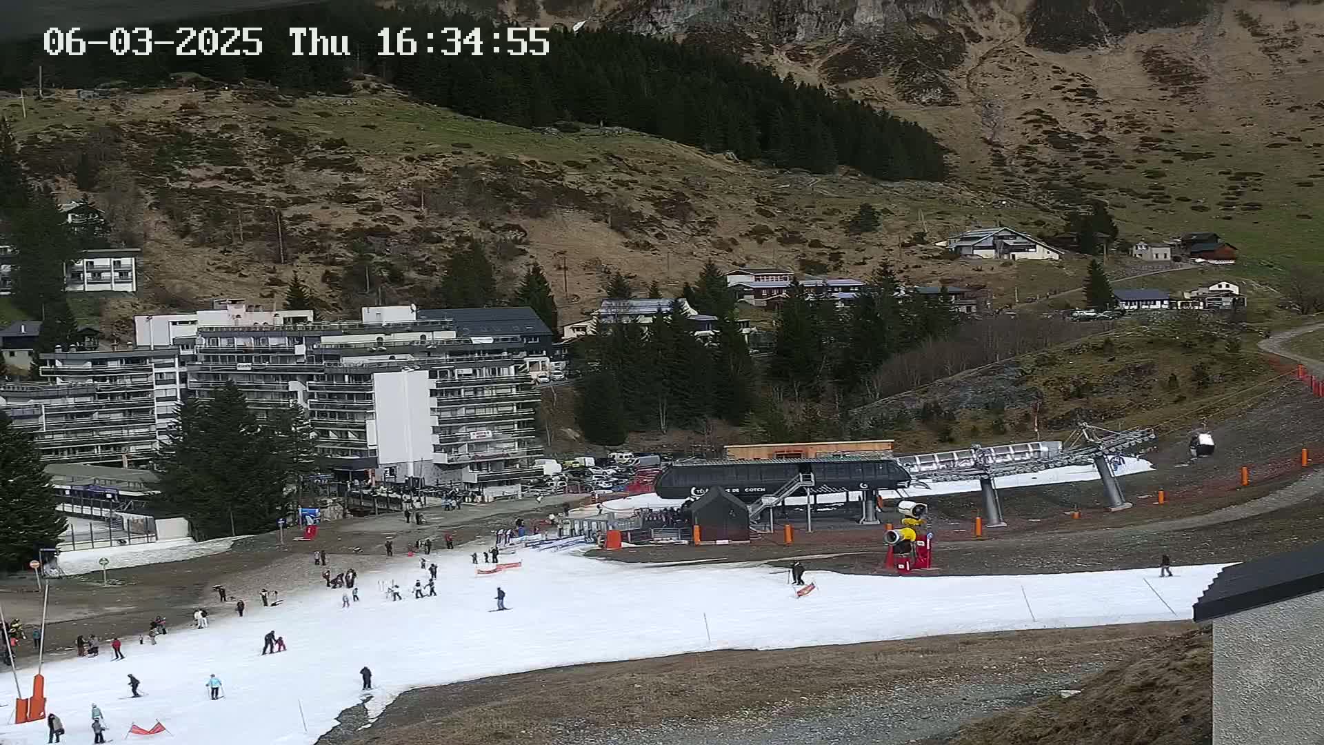 A ski slope with a few skiers, a chairlift, and several buildings nestled in a mountainside under a cloudy sky.