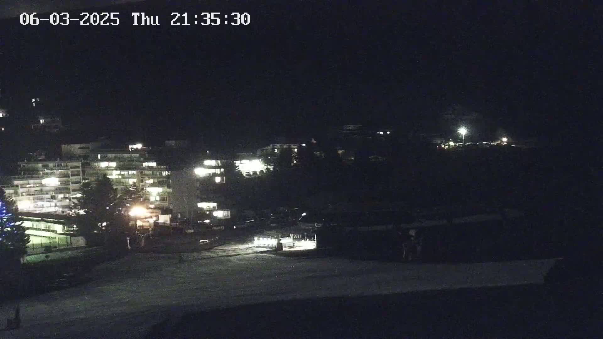 A nighttime view of snow-covered ground in front of several illuminated buildings on a hill.