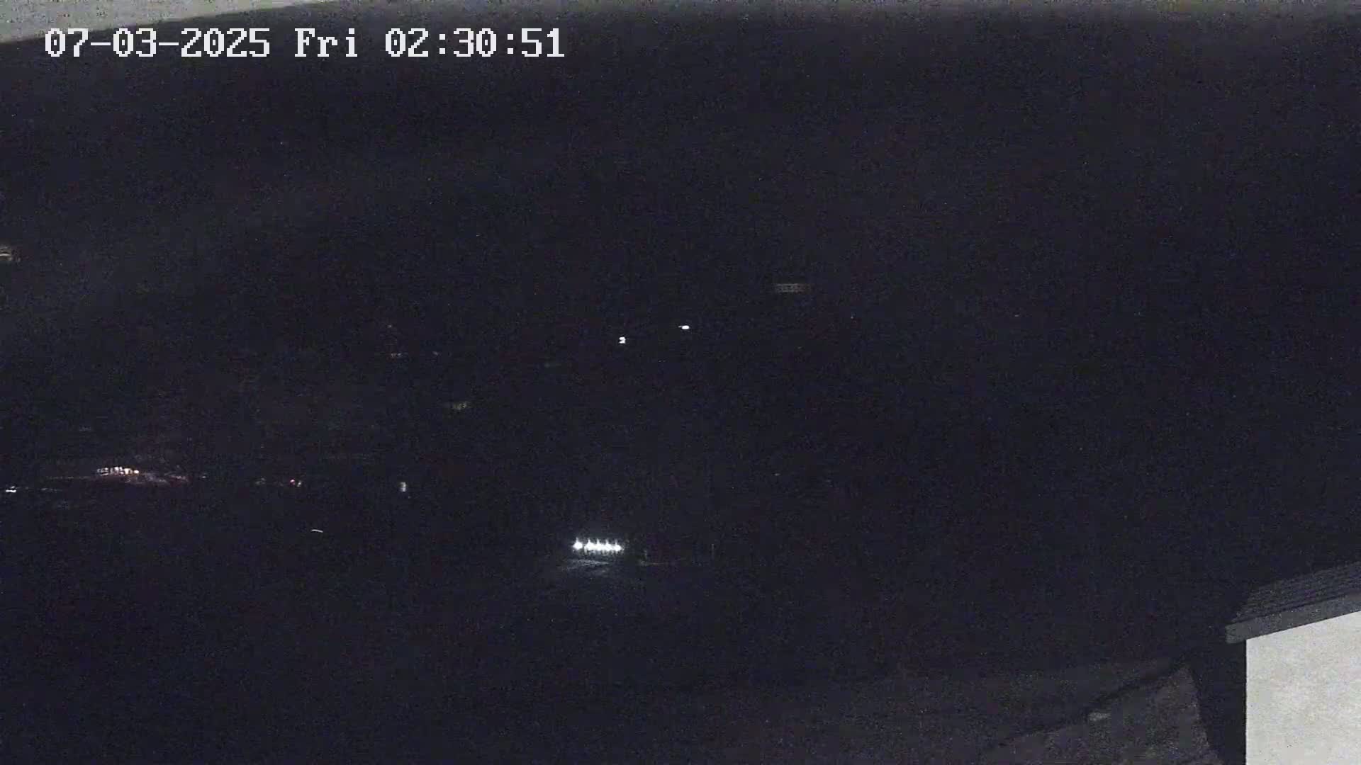 A nighttime, low-light outdoor scene shows a few faint lights in the distance and a corner of a building in the lower right.