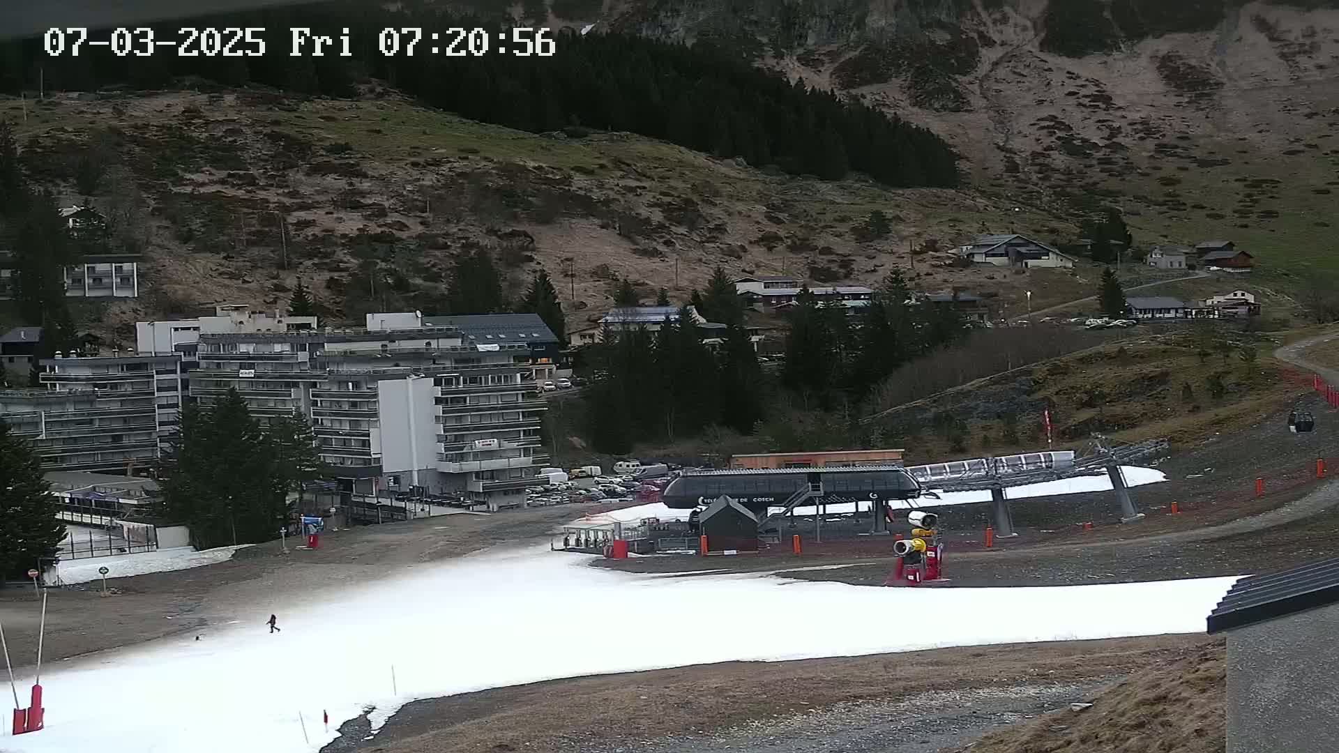 A snow-covered ski slope with a chairlift and a multi-story building sits below a hillside covered in sparse vegetation under an overcast sky.