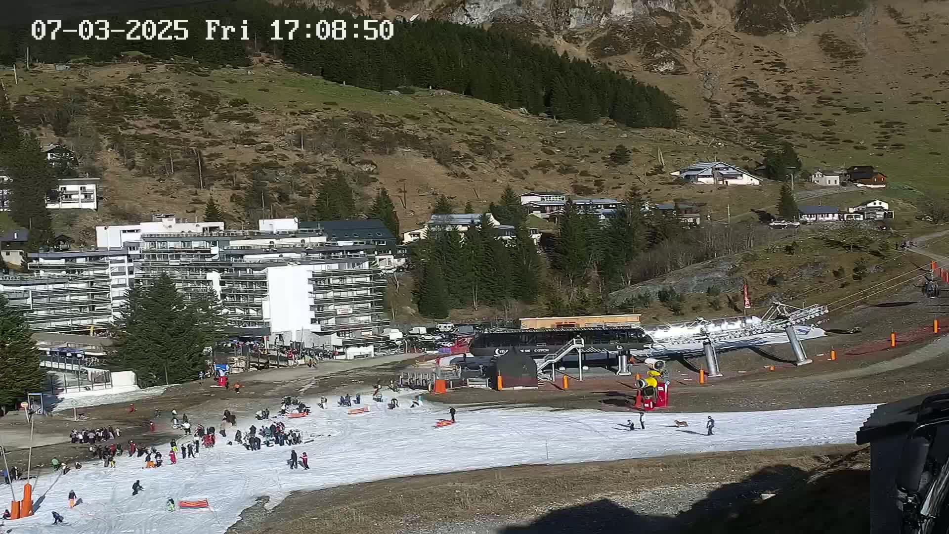 A sunny mountainside ski resort scene shows a snow-covered slope with skiers and snowboarders, a chairlift, buildings, and surrounding trees.