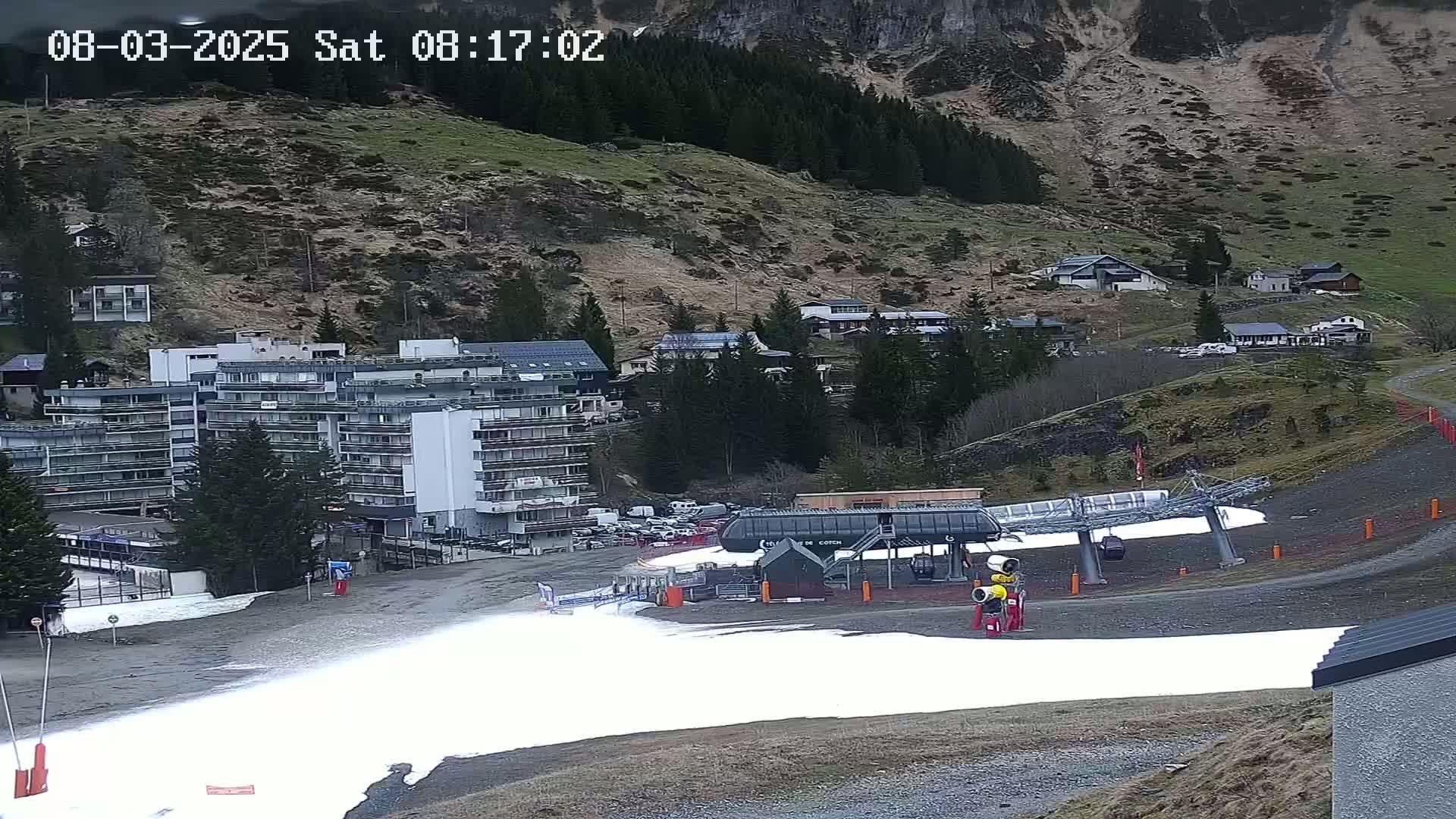A ski lift base with patches of snow sits in front of a large apartment building and several smaller buildings nestled in a mountainside valley under an overcast sky.