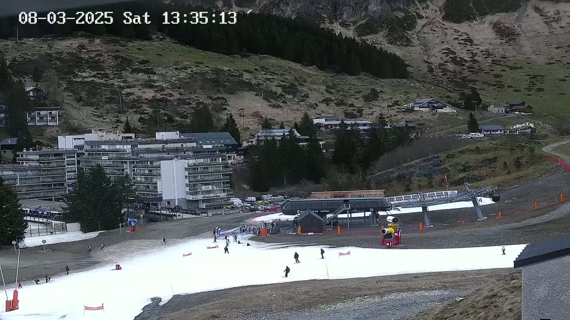 A ski slope with a small amount of snow, a chairlift, and a multi-story building is visible, with a mountainside covered in sparse vegetation in the background under an overcast sky.