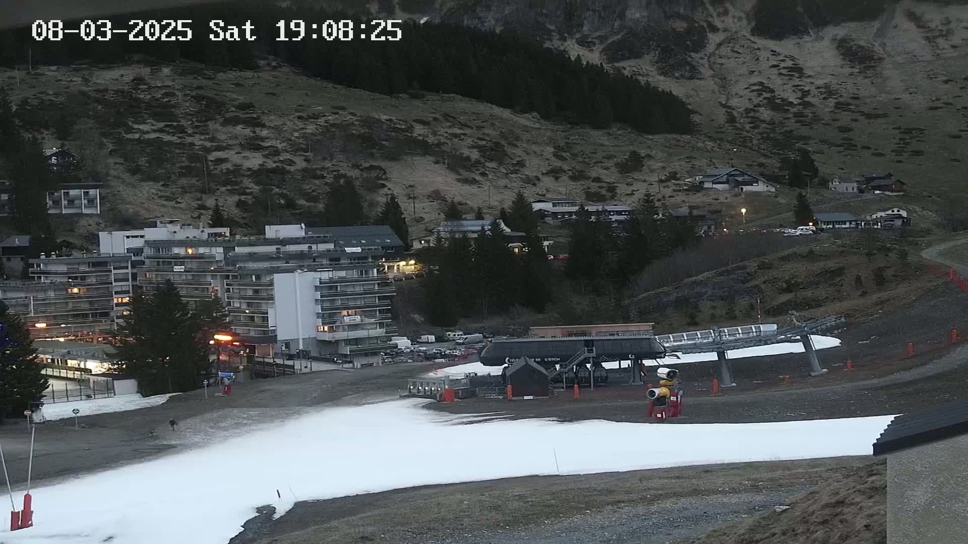 A ski lift sits at the base of a mountainside village under a dark sky, with a patch of artificial snow on the ground.