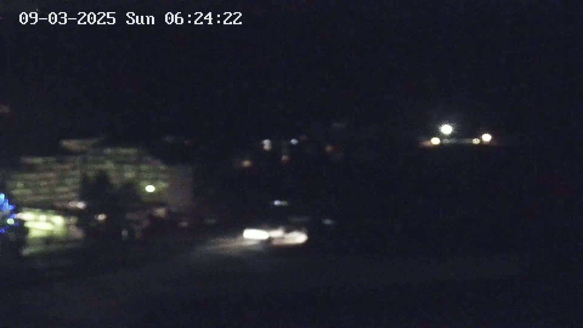 A blurry nighttime image shows a building with lights on in the distance, a vehicle's headlights in the mid-ground, and a bright moon in the sky.