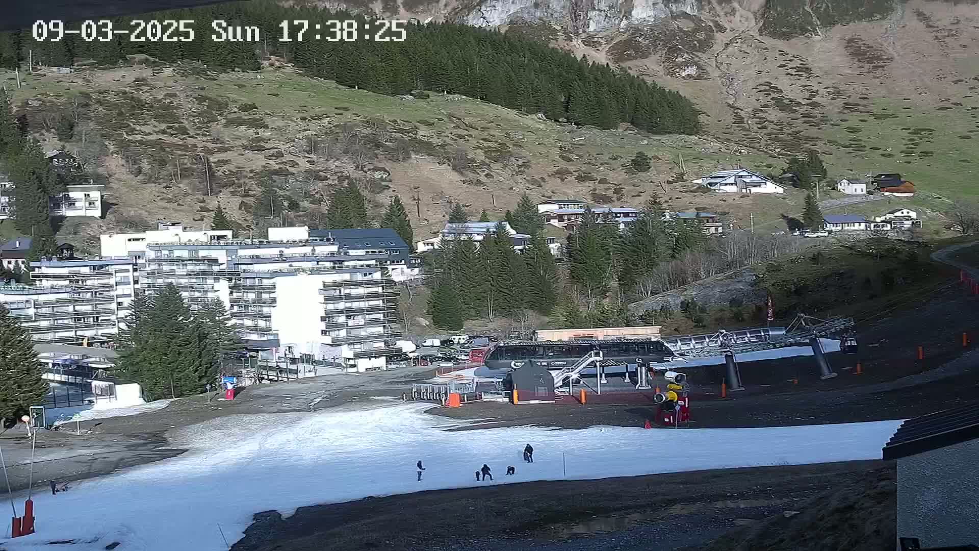 A sunny mountainside village with a large apartment building, a ski lift, and patches of snow on the ground.
