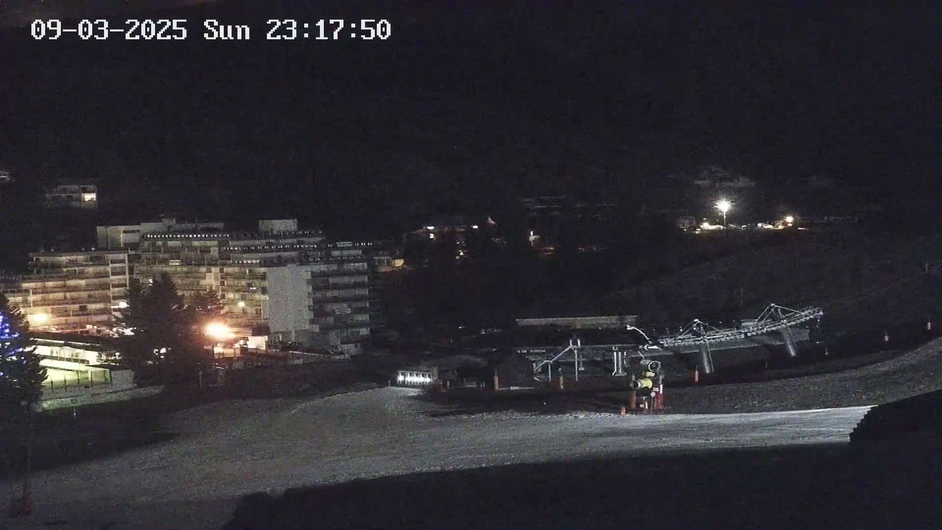 A nighttime view of a snow-covered ski slope with a building and ski lift in the background under dark conditions.