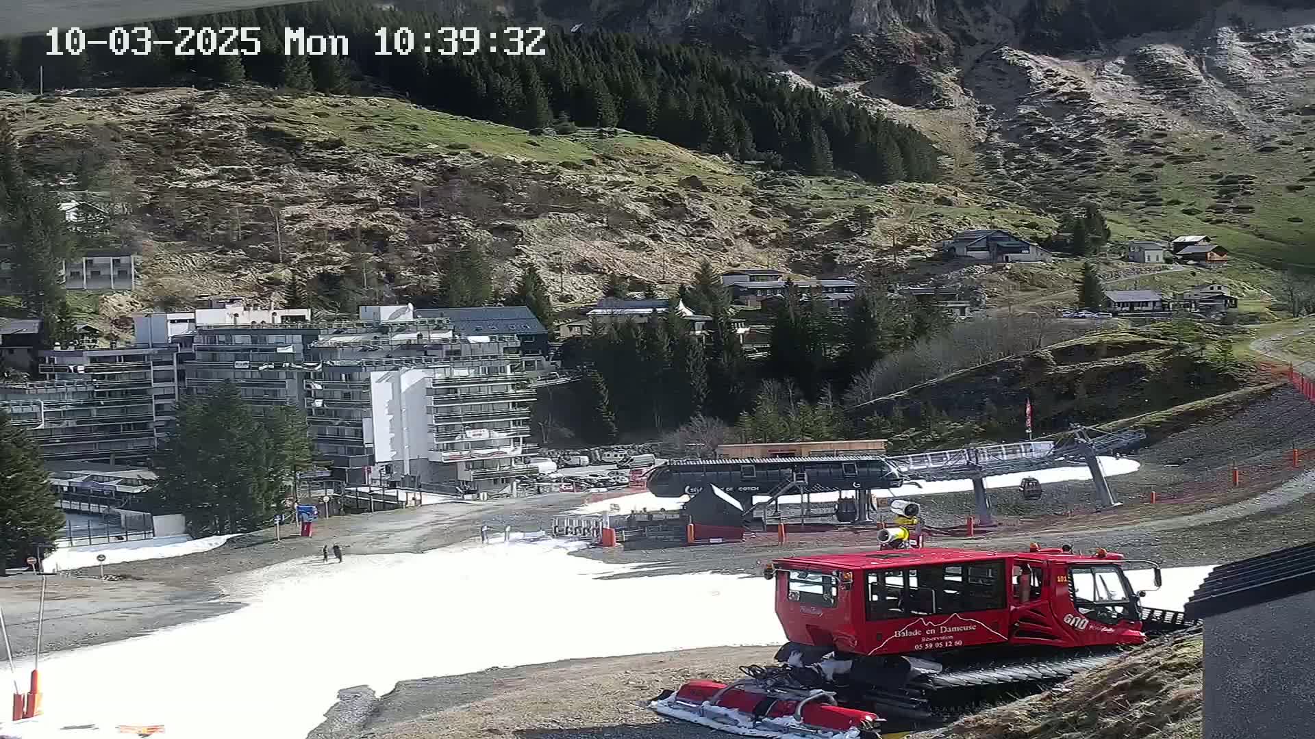 A red snow groomer sits on a snow-covered area below a multi-story building and ski lift, nestled in a mountainous region under sunny conditions.