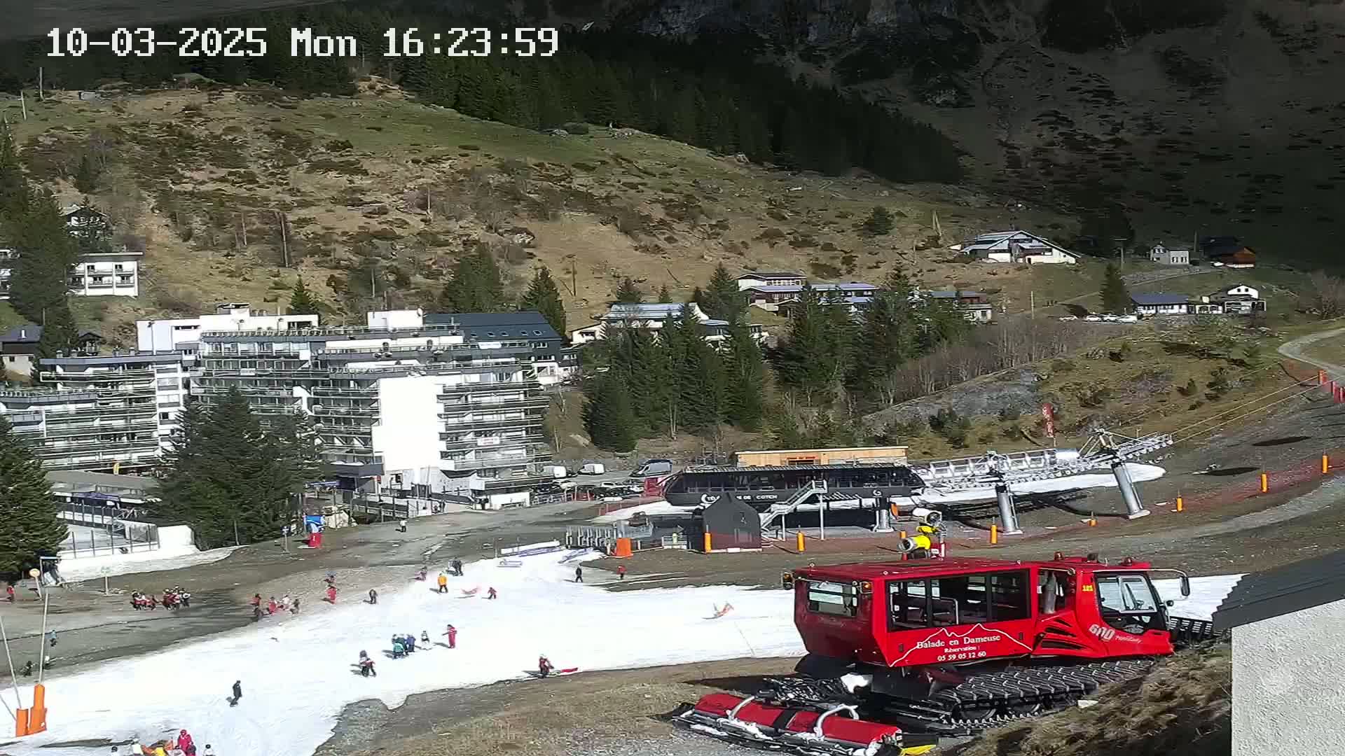 A snowcat sits on a mostly snow-free ski slope near a multi-story building and several smaller buildings, all nestled in a sunny mountainside.