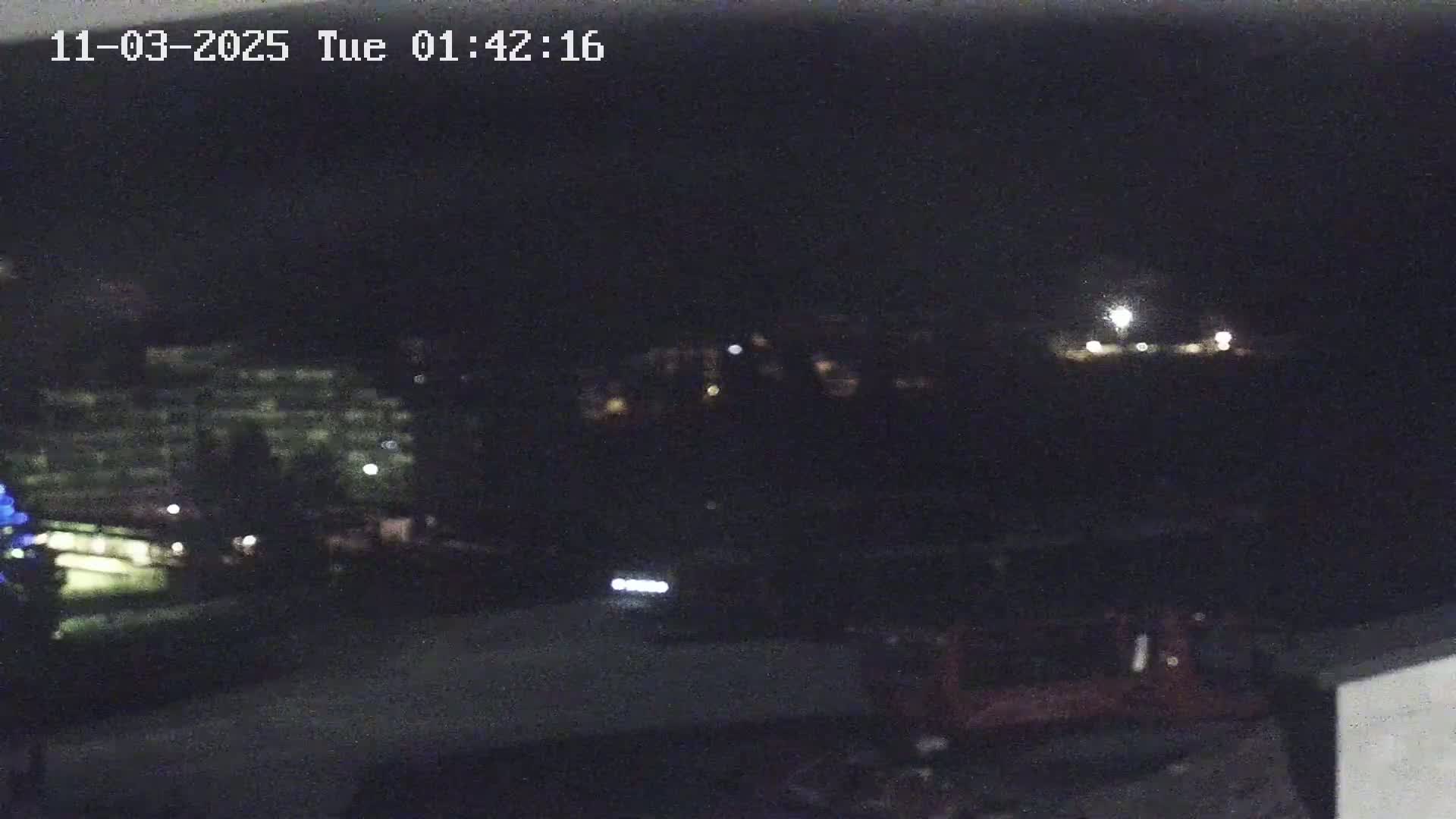 A nighttime, blurry view of dimly lit buildings and a road with a vehicle's headlights visible in the distance.