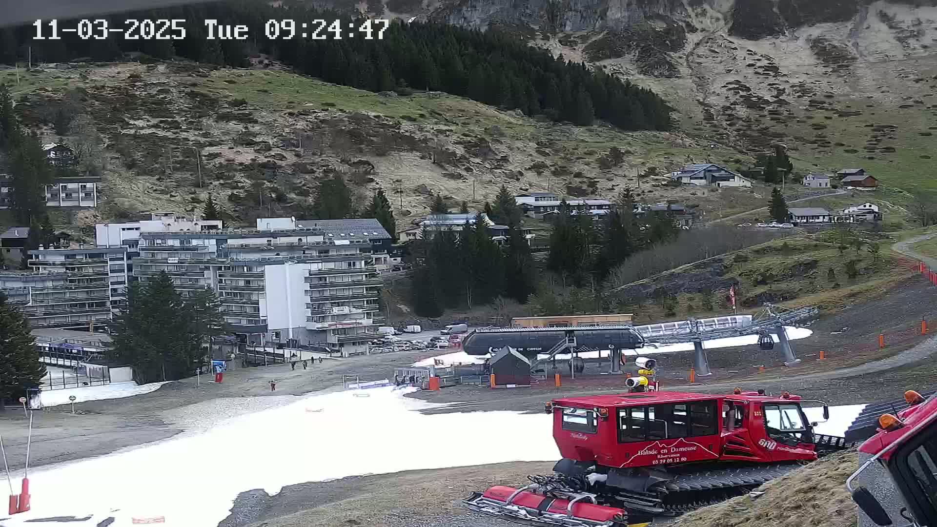 A red snowcat is parked near a ski lift and a large apartment building in a mountainous area with patchy snow on the ground.