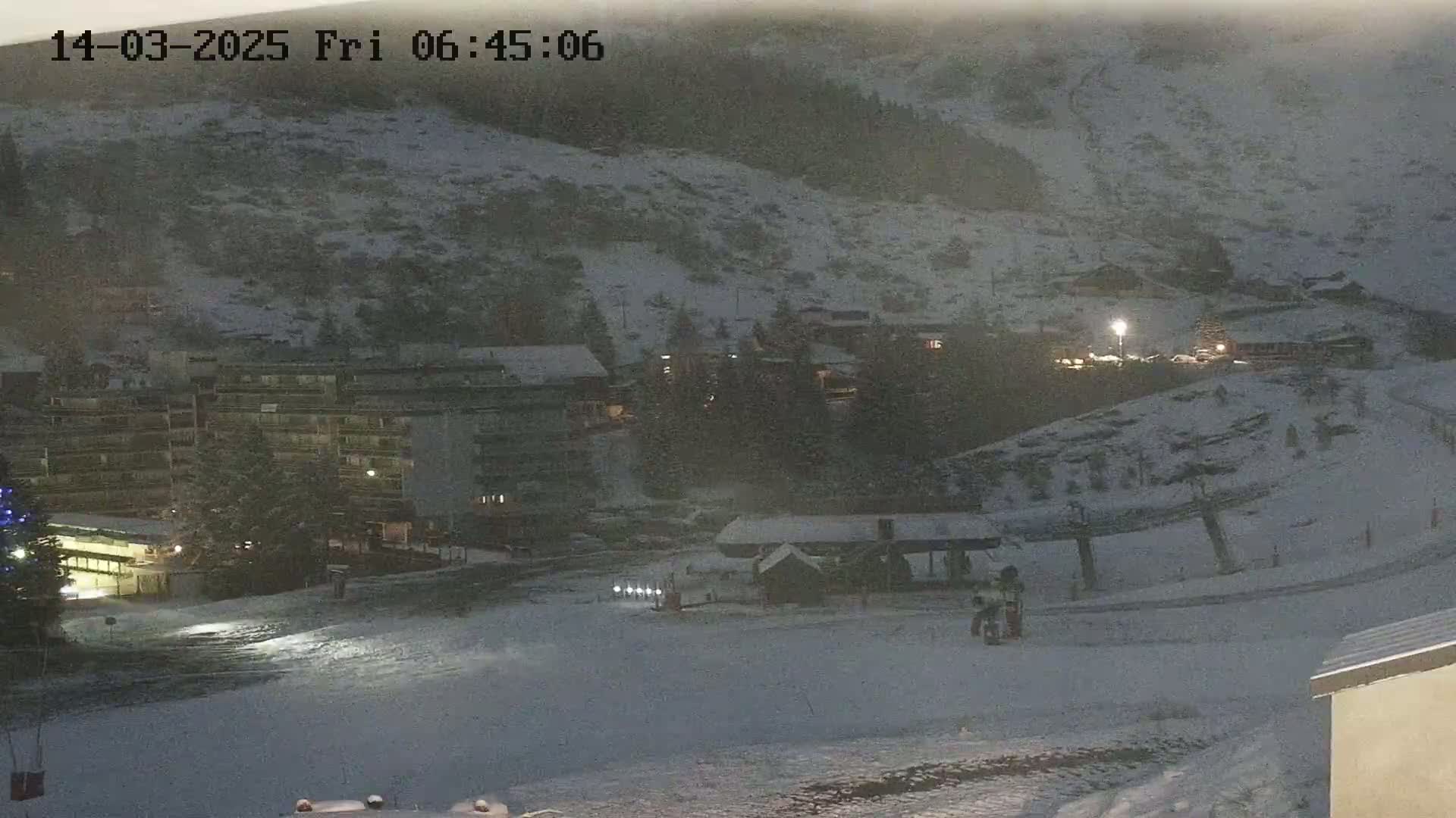 A snow-covered mountainside village is visible at night under falling snow, with buildings and a ski lift illuminated.
