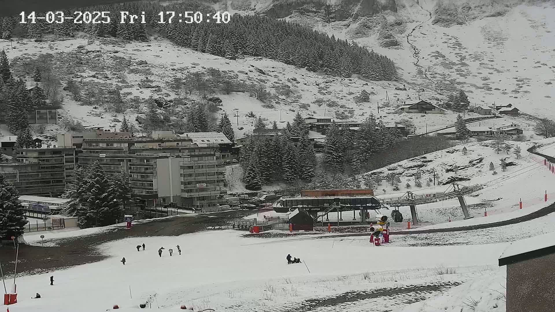 A snow-covered ski resort with a chairlift, several buildings, and a few people scattered across a snowy slope under a light snowfall.