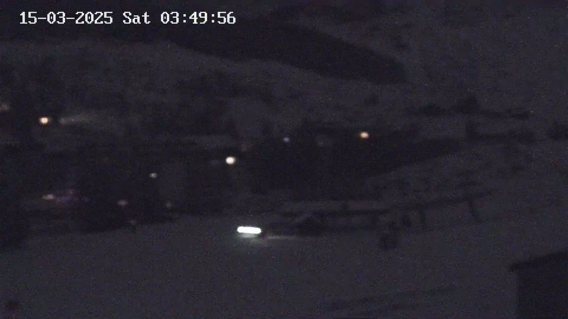 A nighttime, low-light image shows a dark, snow-covered area with a few distant lights and a bright headlight in the lower center.
