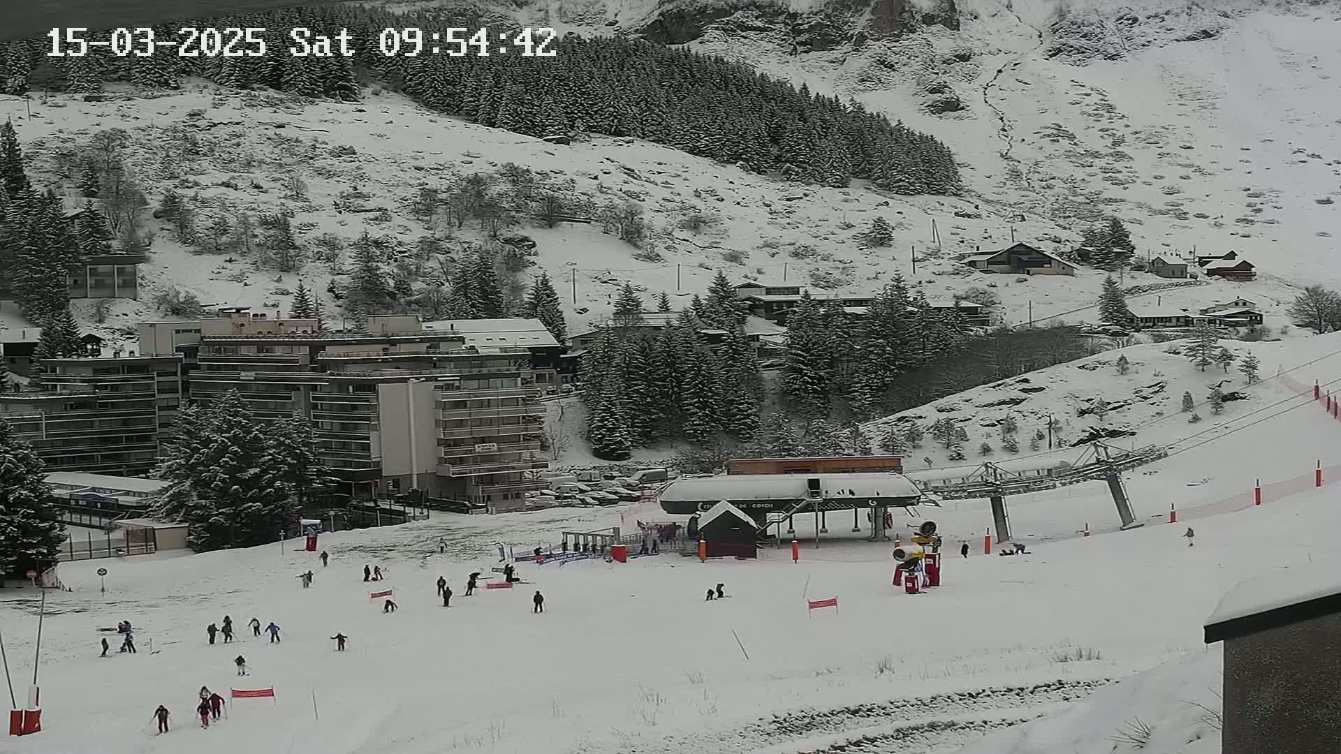A snowy ski slope with several skiers, a ski lift, and buildings nestled in a snow-covered mountainside.