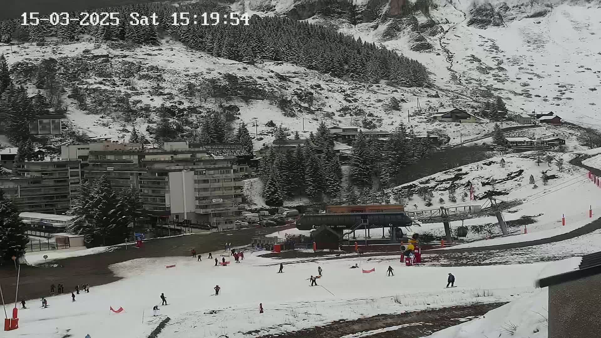 A snow-covered ski slope with several skiers and a ski lift is situated in front of a snow-dusted mountainside with buildings and evergreen trees.
