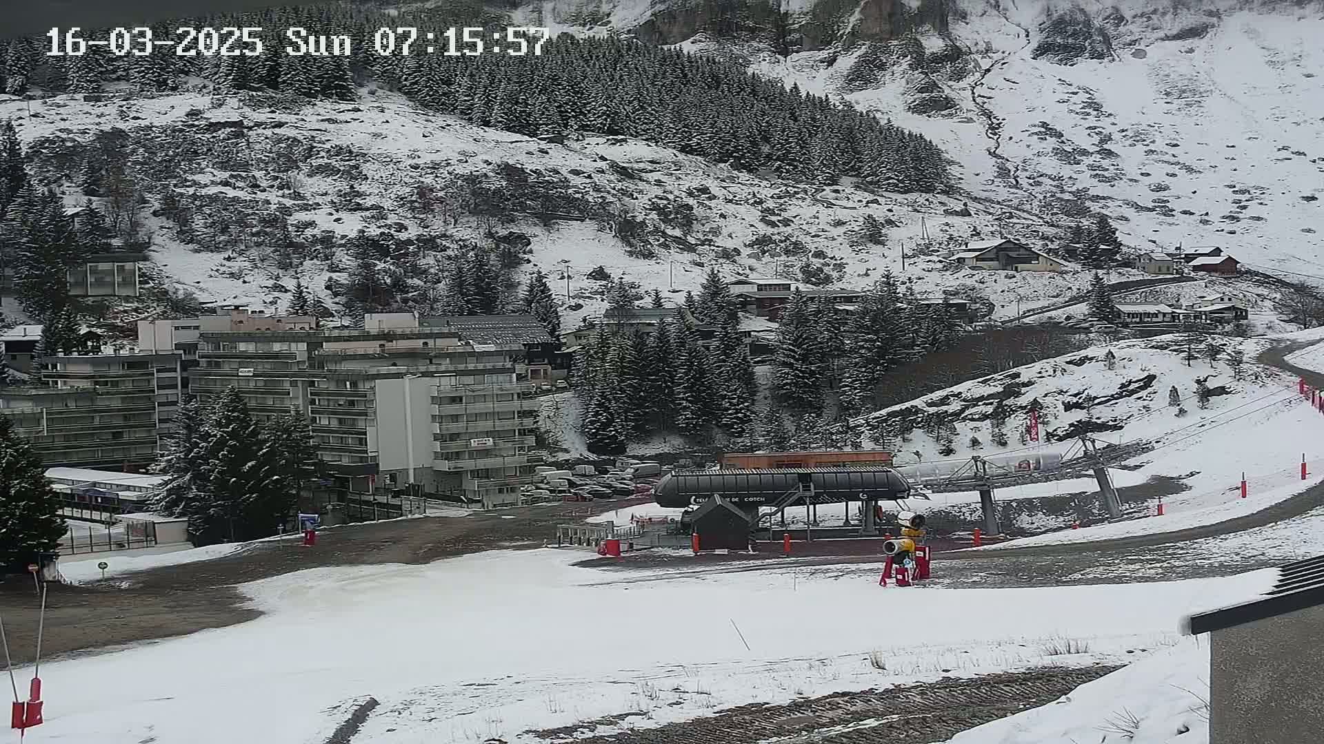 A snow-covered ski resort with a chairlift, buildings, and a mountain in the background under snowy conditions.