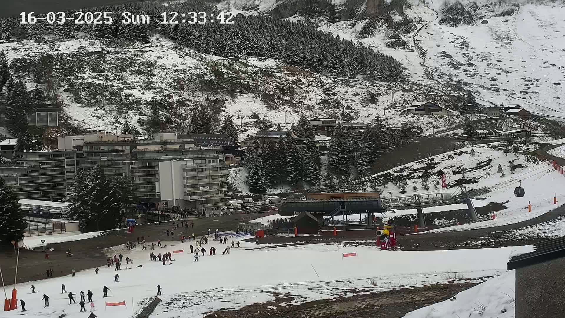A snow-covered ski resort with several buildings, a chairlift, and numerous skiers and snowboarders on the slopes under an overcast sky.