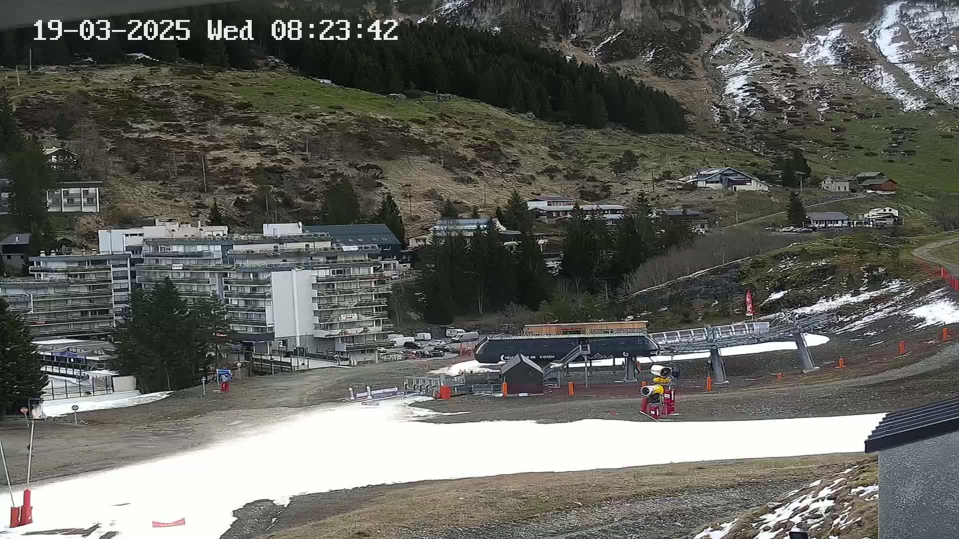 A ski lift base station sits at the bottom of a mountainside with patches of snow, near a multi-story building and some smaller residential structures; the mountainside is mostly barren with some trees and patches of snow.