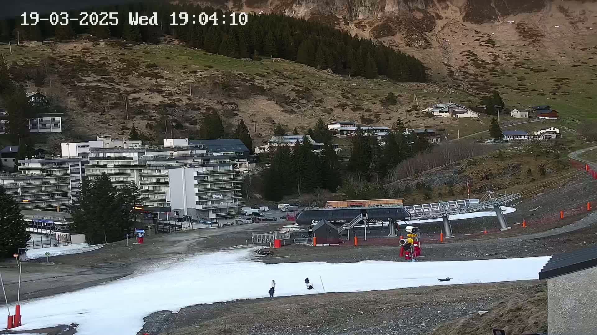 A ski lift and a small amount of snow are visible in a mountain village, with buildings and a hillside in the background under twilight conditions.