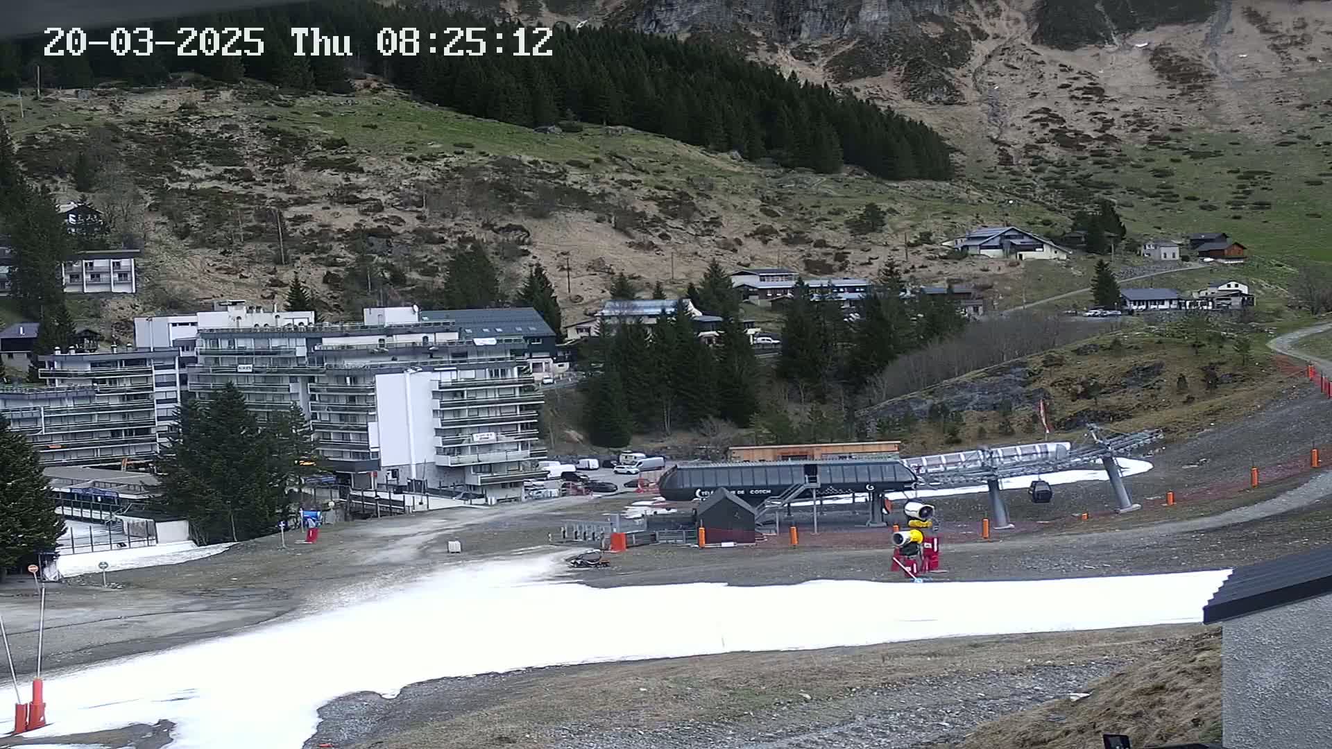 A ski lift base station with a patch of snow sits below a hillside village nestled in a mountainside under an overcast sky.